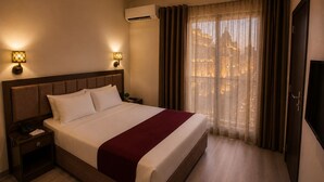 A hotel room with a large bed, two bedside lamps, an air conditioning unit, a TV, and a window with curtains.