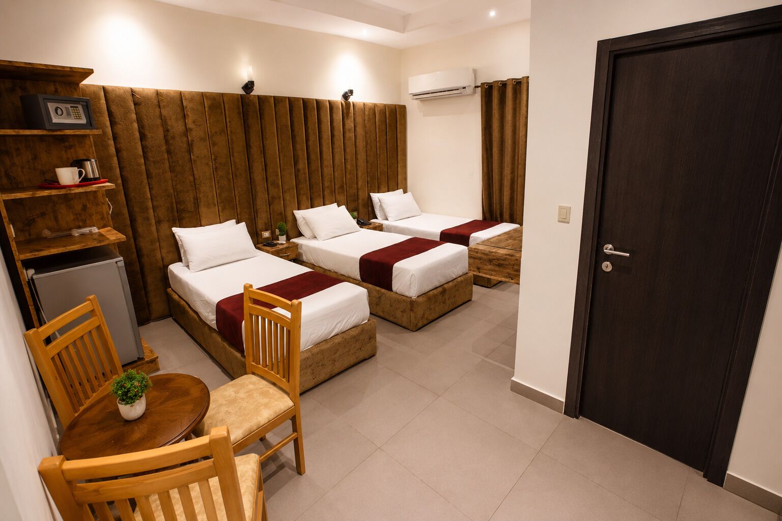 A hotel room with two beds, a small table with chairs, a plant, a refrigerator, and a door.