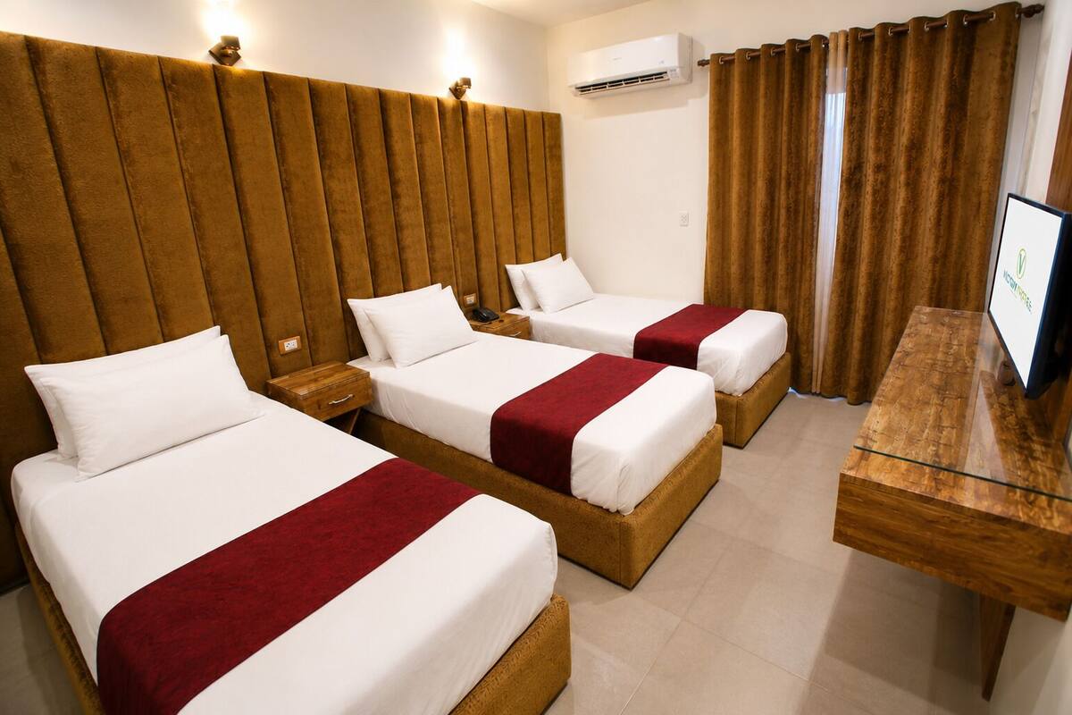 Habitación de hotel con tres camas, cabecera de madera, televisión y aire acondicionado.
