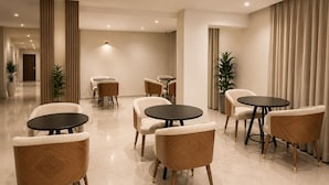 A modern hotel lobby with a marble floor, several round tables with chairs, and a large plant.