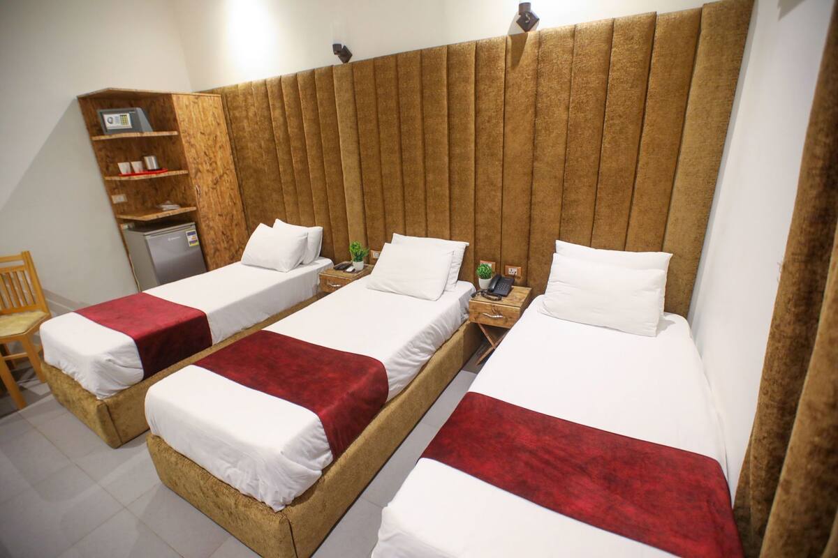 Una habitación con tres camas individuales, un cabecero de madera, un pequeño escritorio con teléfono y un estante con microondas.
