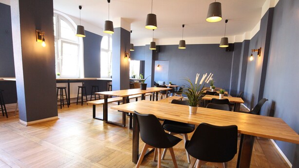 A modern dining area with wooden tables and black chairs, featuring pendant lighting and potted plants. - Blue Doors Hostel KTV (Rostock)