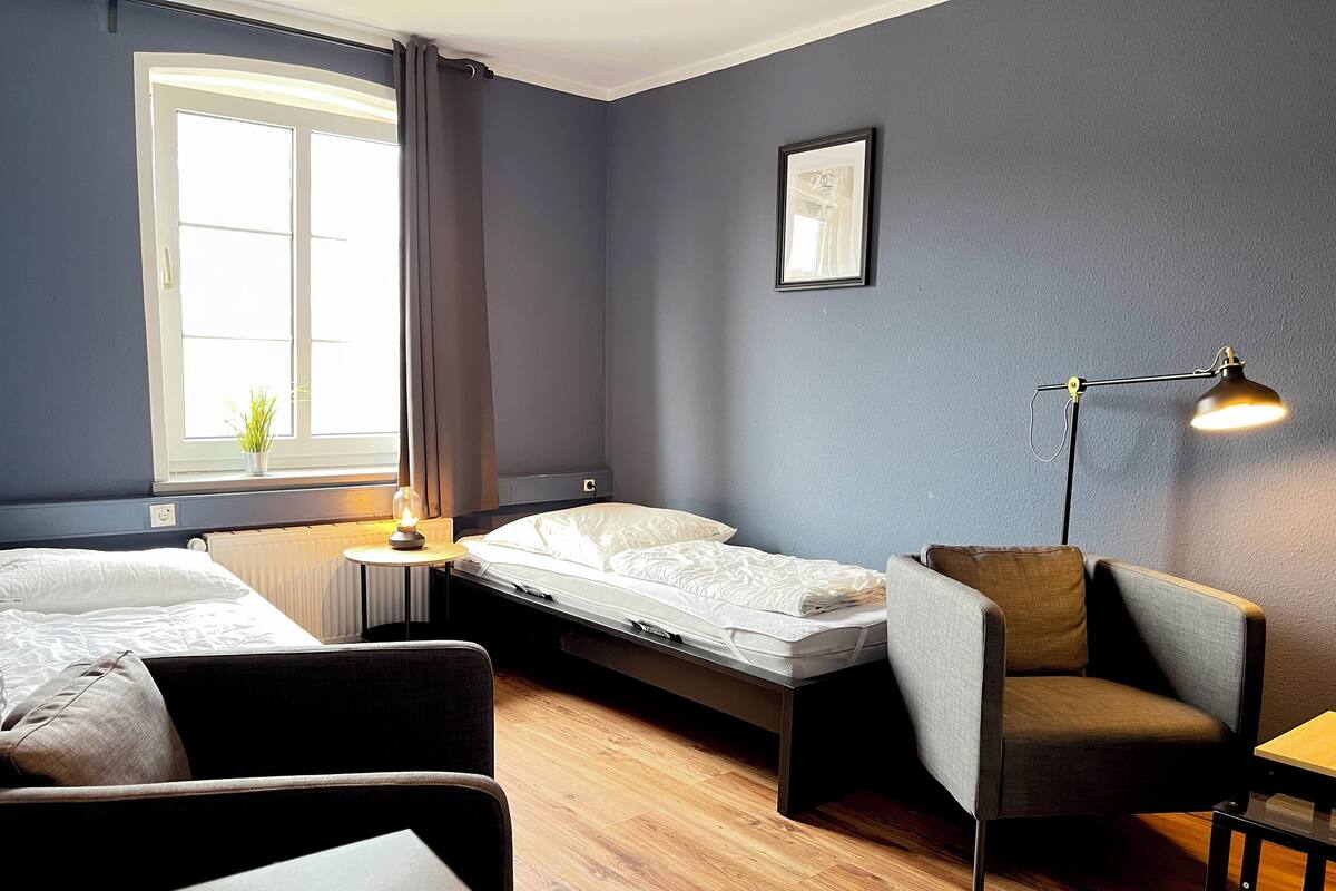 A hotel room with two beds, a chair, a table, a lamp, a window with curtains, a radiator, and a framed picture on the wall.