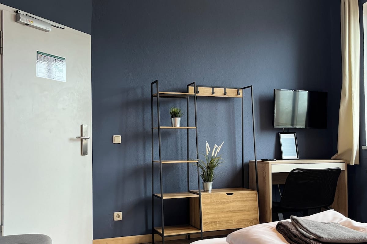 A bedroom with a bed, a desk, a chair, a TV, and a shelf with a plant.