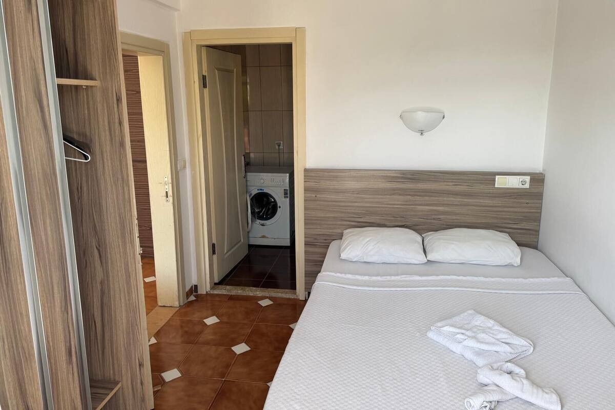 Une chambre d’hôtel avec une armoire en bois, un lit avec du linge de lit blanc, un lave-linge dans la pièce adjacente et un luminaire fixé au mur.