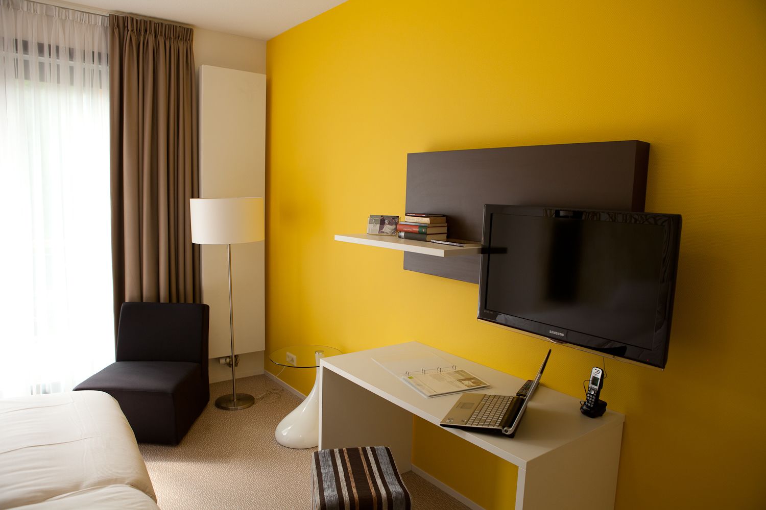 Ein Hotelzimmer mit gelber Wand, einem Flachbildfernseher, einem Schreibtisch mit einem Laptop, einem Sessel, einem Nachttisch und einer Lampe.