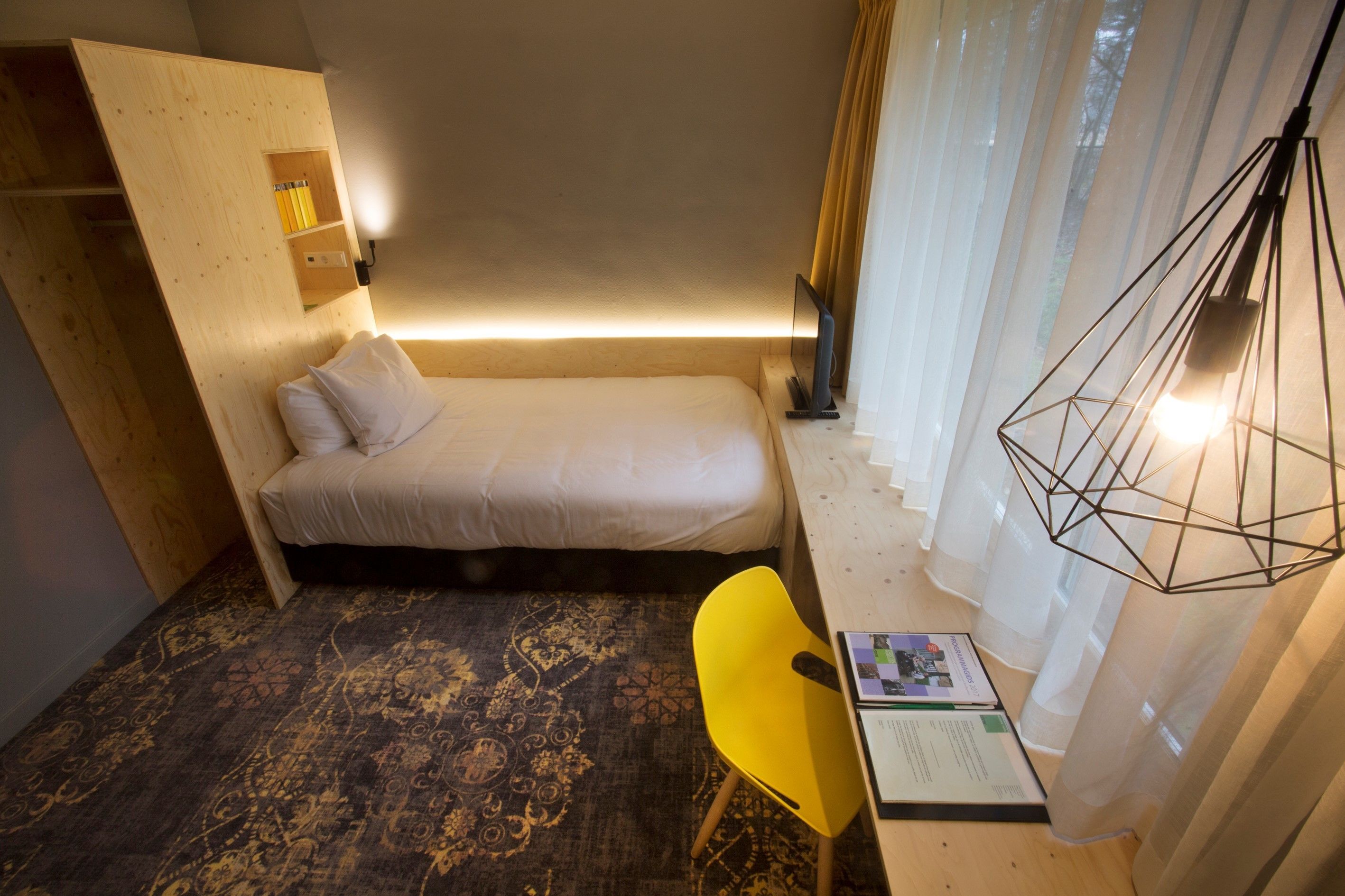 A compact bedroom with a bed, a desk with a chair, and a window with sheer curtains.