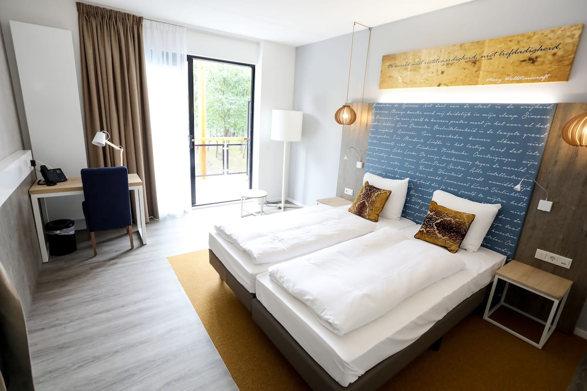 A hotel room with two beds, a desk, a chair, a lamp, and a view of the outdoors.