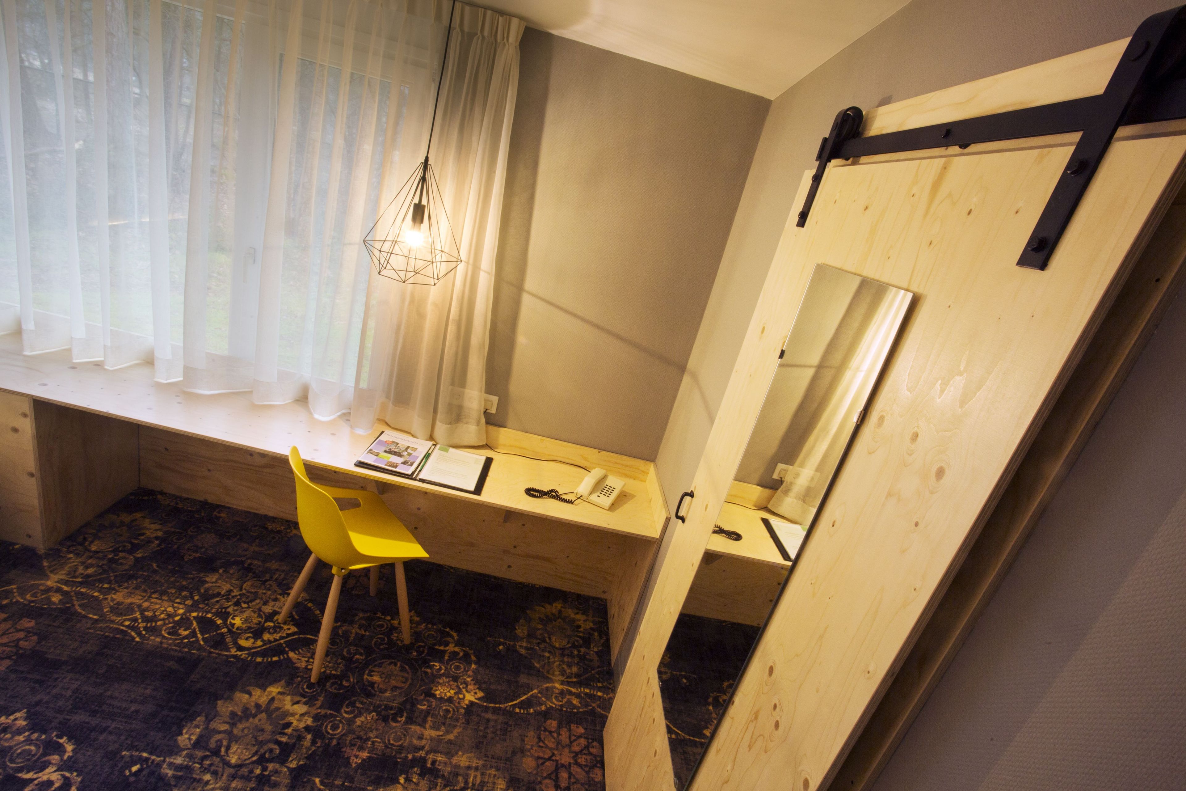 A compact room with a wooden desk, a yellow chair, a large mirror, and a window with sheer curtains.