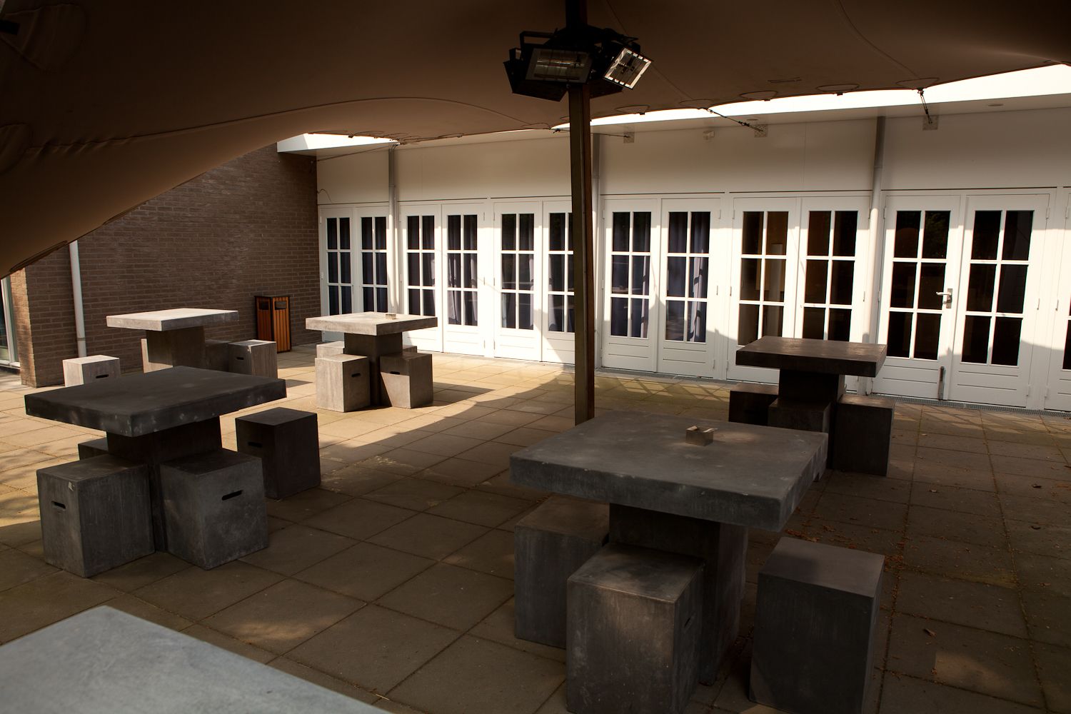 An outdoor patio area with multiple concrete tables and benches, a brick wall, and large windows with white frames.