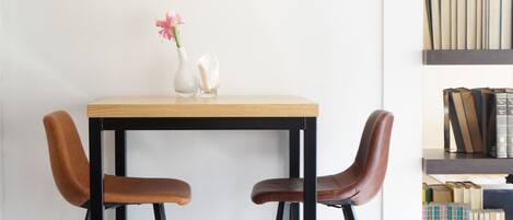 A minimalist room with a wooden table, two leather chairs, a black pendant light, a white vase with pink flowers, and a bookshelf with books.