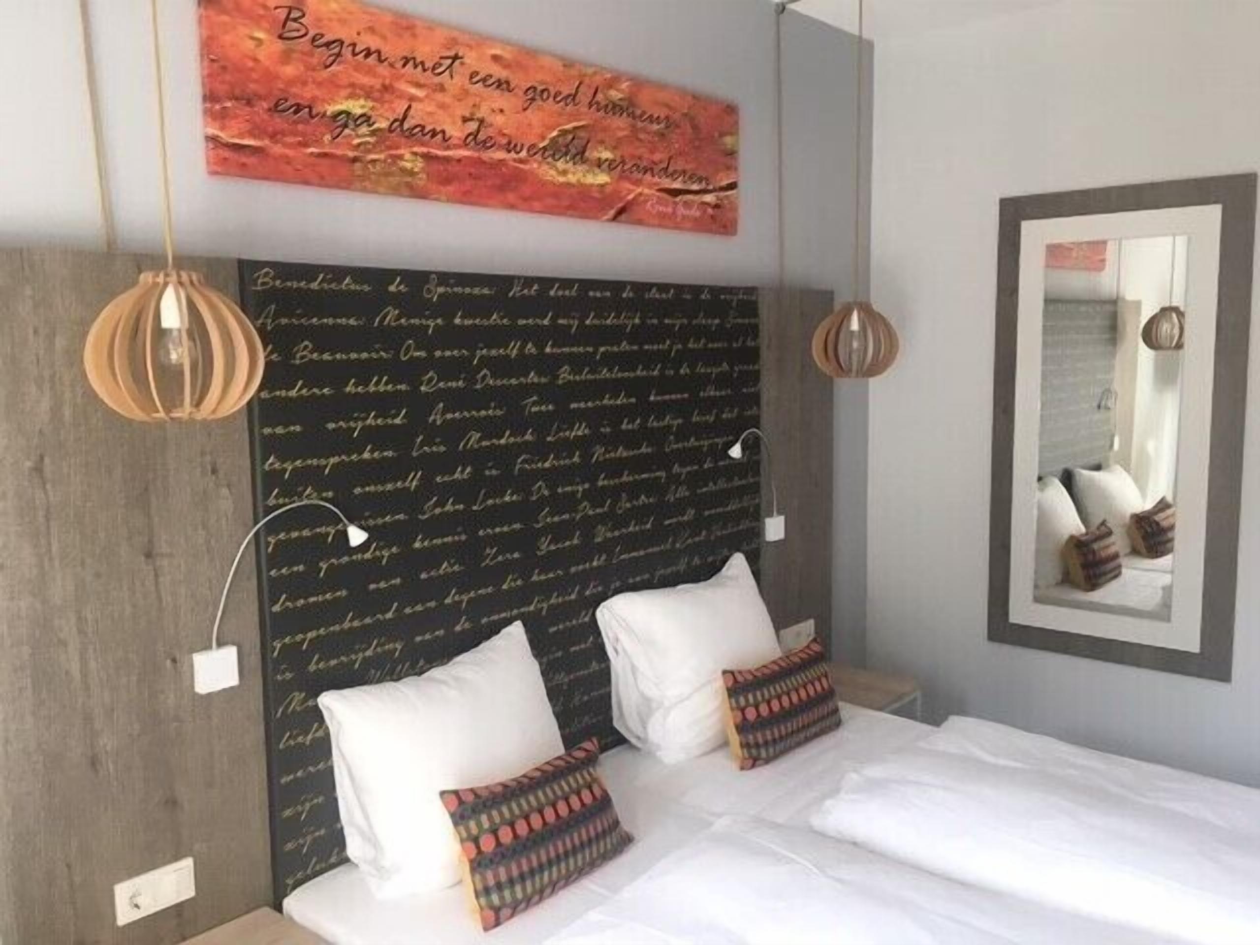 A bedroom with a large bed, two pillows, a wall with a quote, and a mirror.