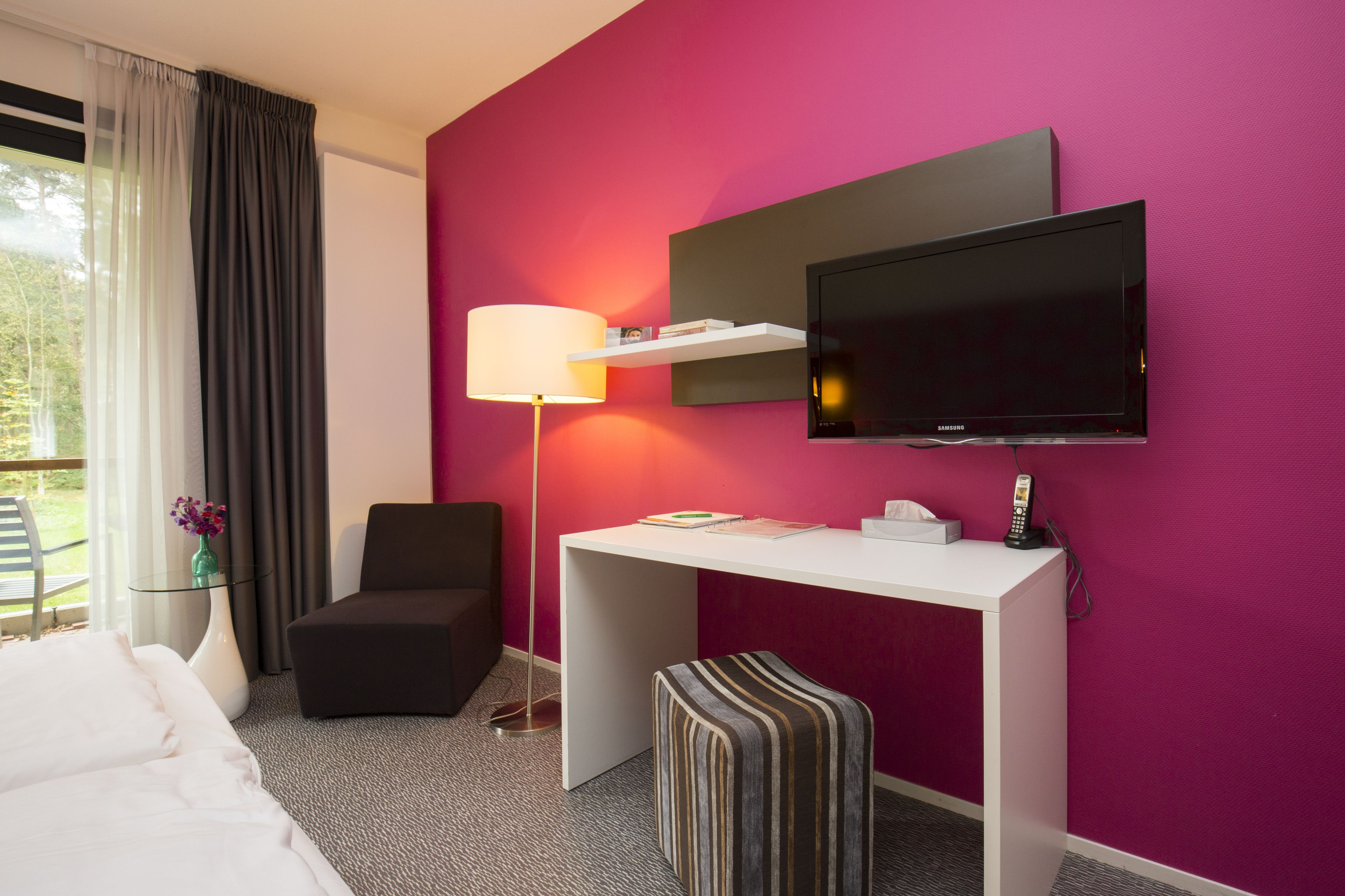 A hotel room with a bed, a desk with a lamp, a TV, a chair, and a small table with a vase of flowers.