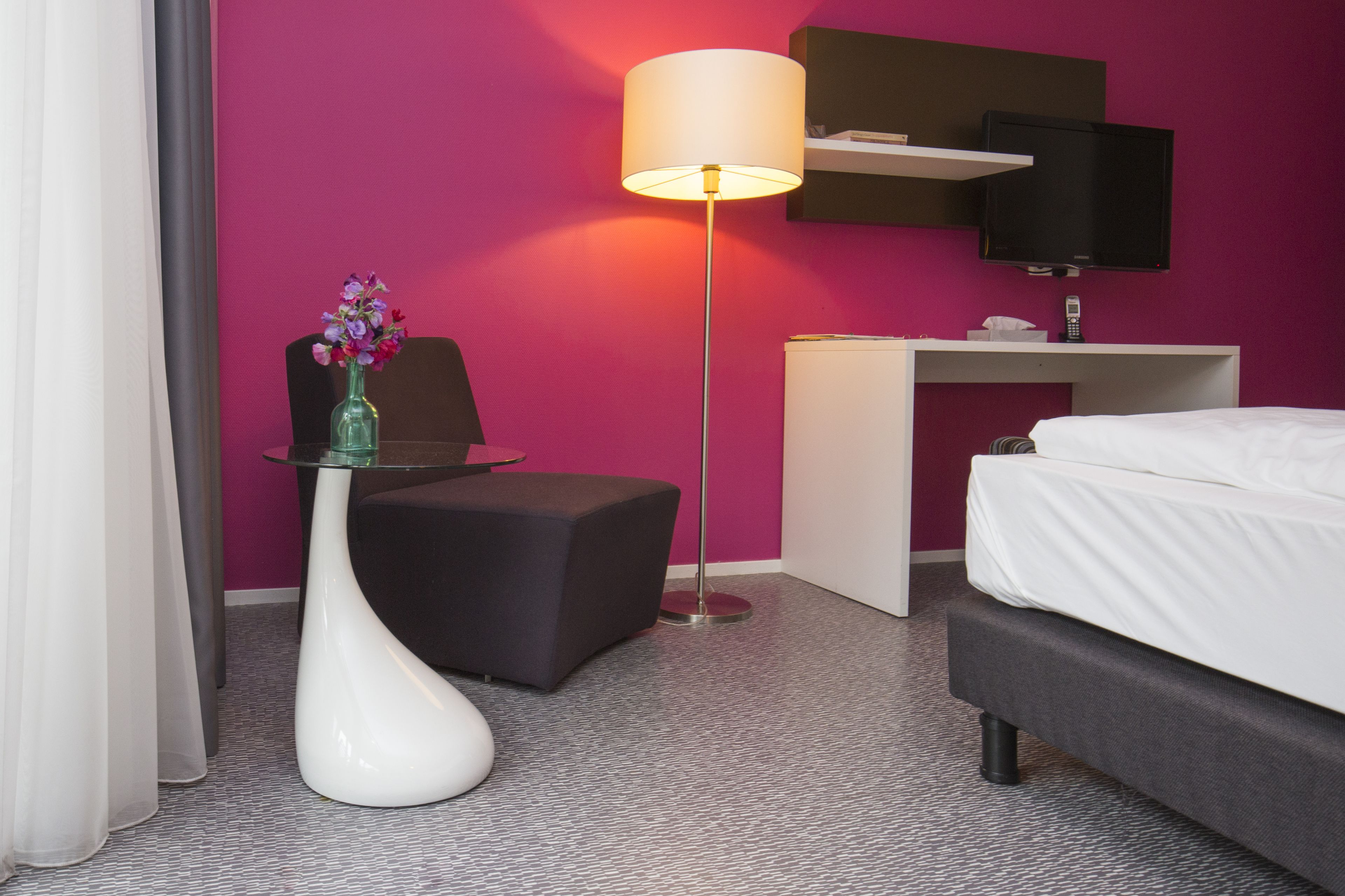 A hotel room with a bed, a desk, a chair, a lamp, and a vase with flowers.