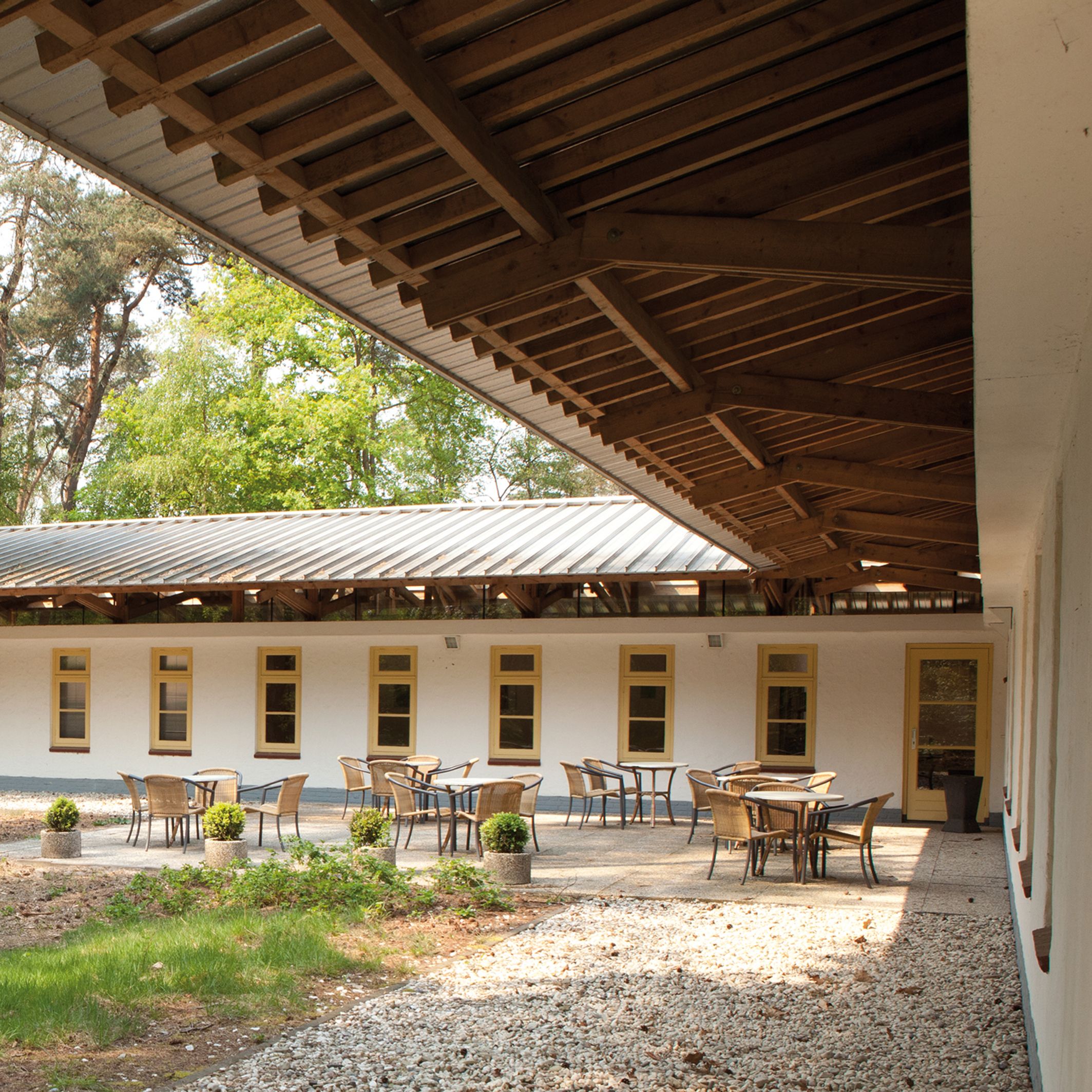 A covered outdoor seating area with tables and chairs, a building with windows, and a gravel path.