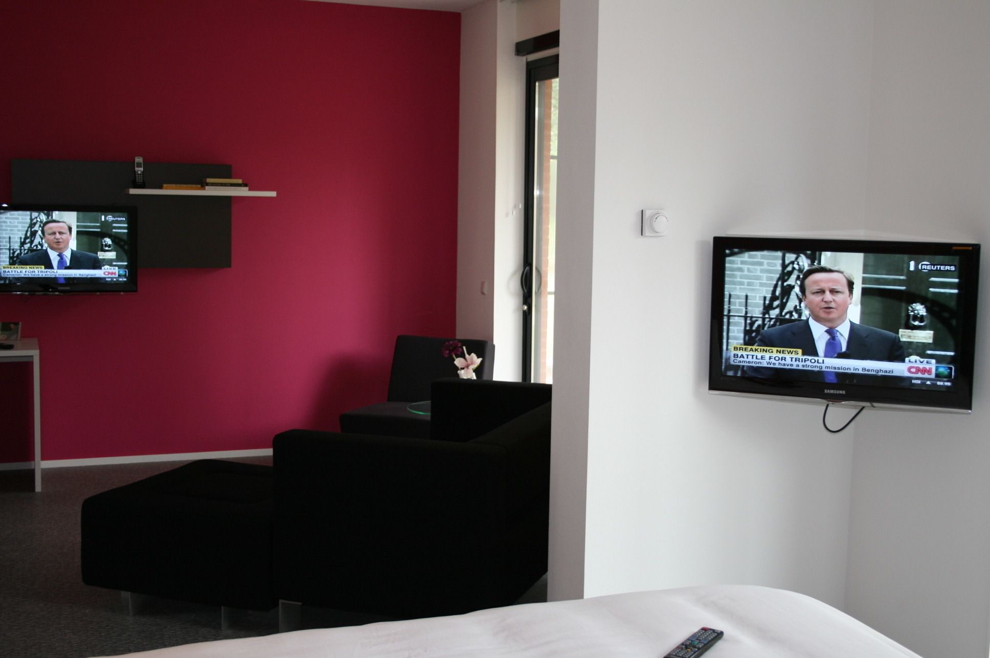 Habitación de hotel con cama, un sillón negro y dos televisores mostrando noticieros.