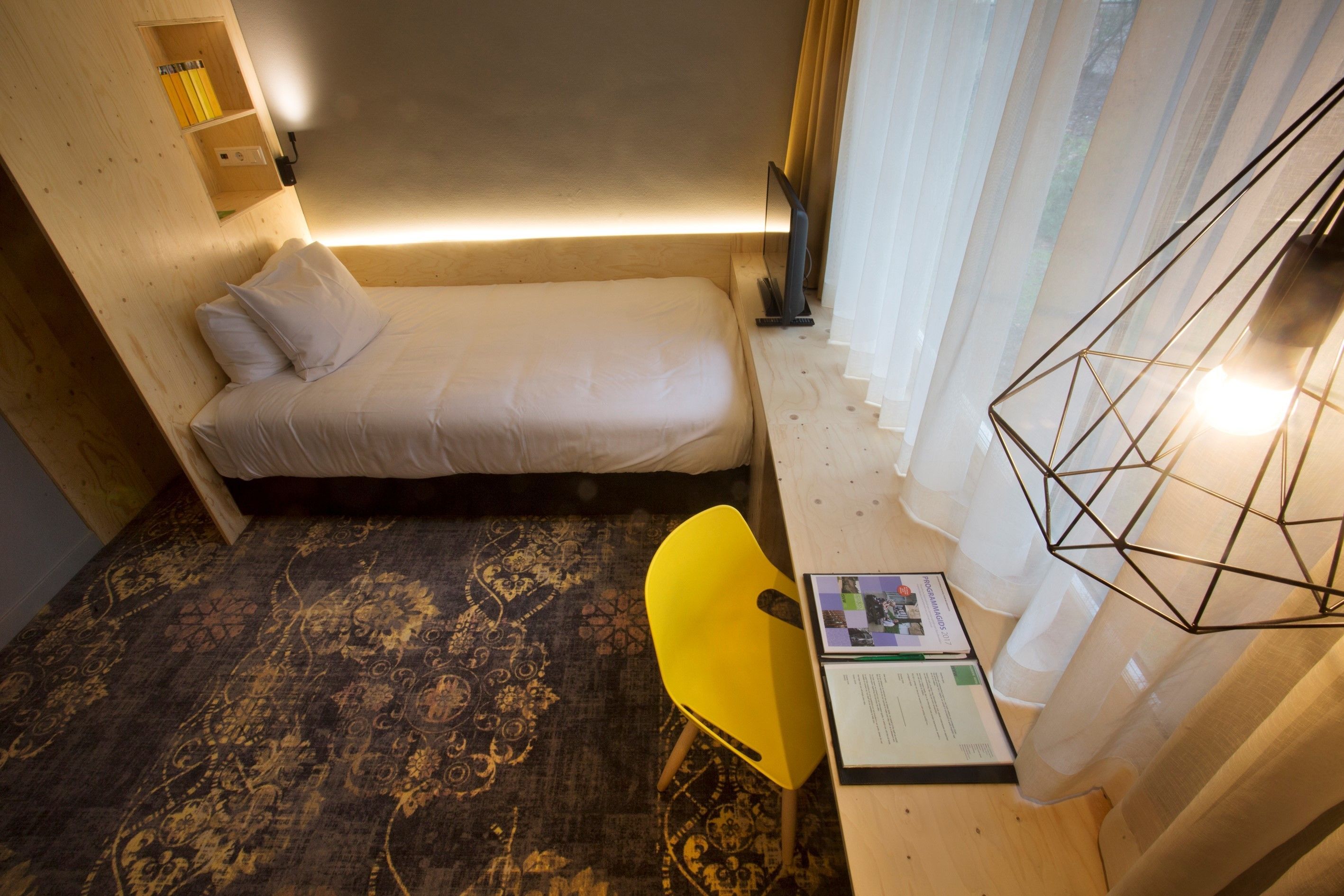 A small, compact room with a single bed, a desk, and a chair. There is a lamp on the desk and a window with curtains.