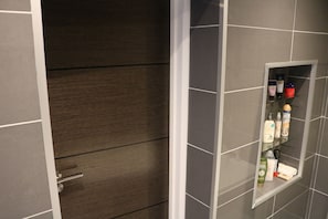 A bathroom with grey tiles, a wooden door, and a glass shelf with toiletries.