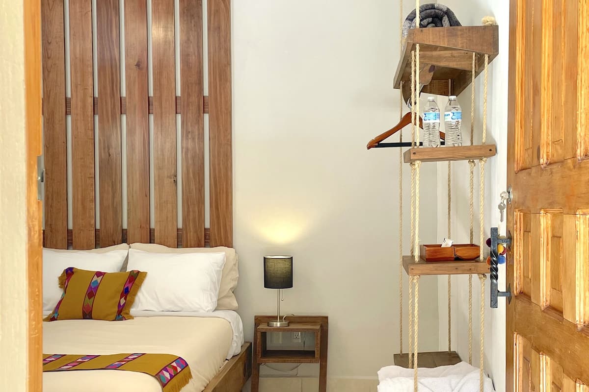Un dormitorio con una cama de madera, una mesita de noche con una lámpara, un armario de madera y un perchero con toallas y artículos de tocador.