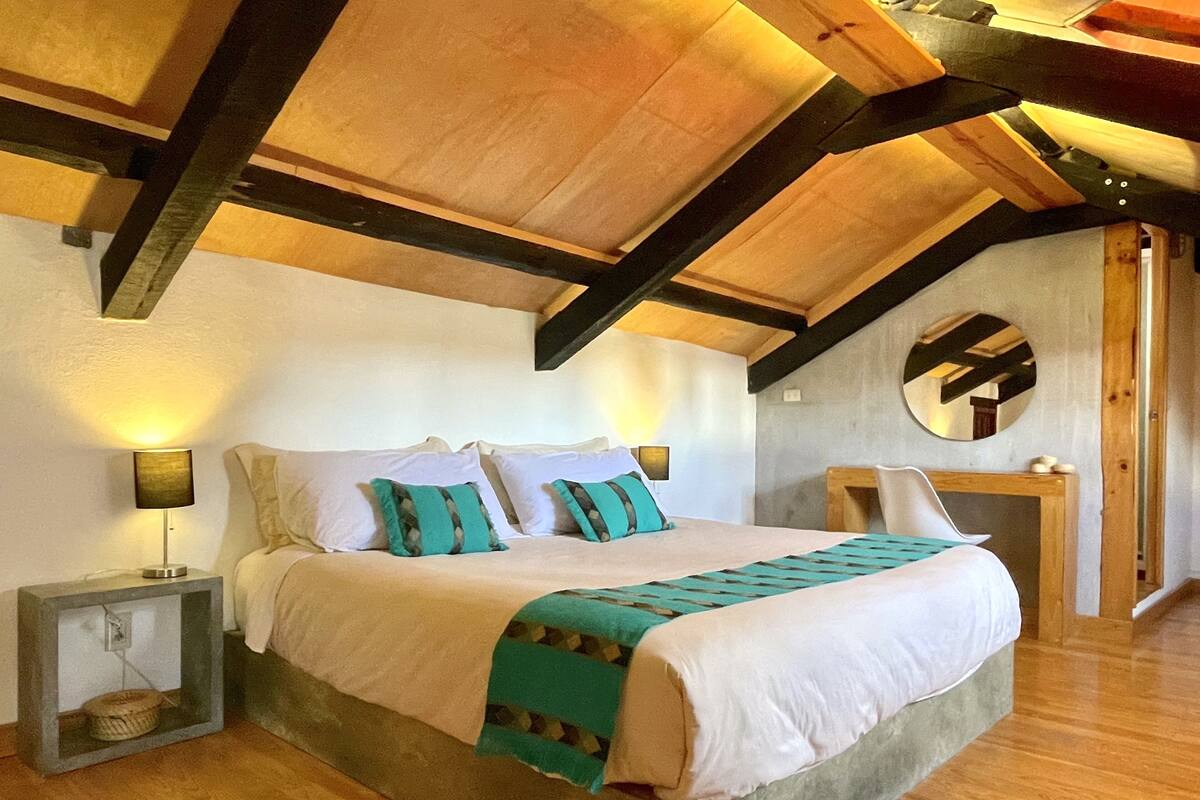 Un dormitorio con una cama grande, vigas de madera, un espejo redondo y una mesita de noche con lámpara.