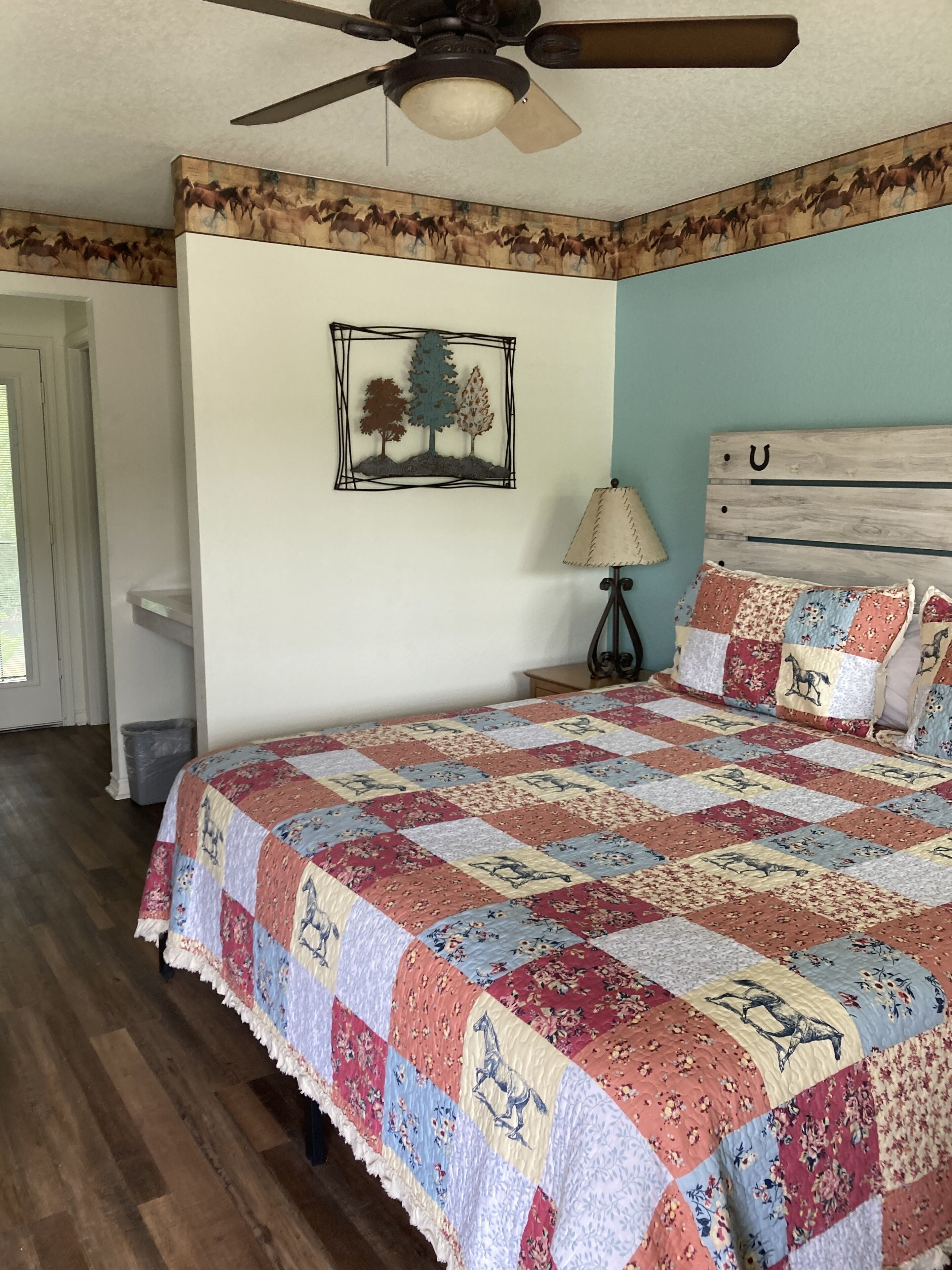 A bedroom with a large bed, a ceiling fan, a wall-mounted picture of trees, and a wooden headboard with a 'U' design.