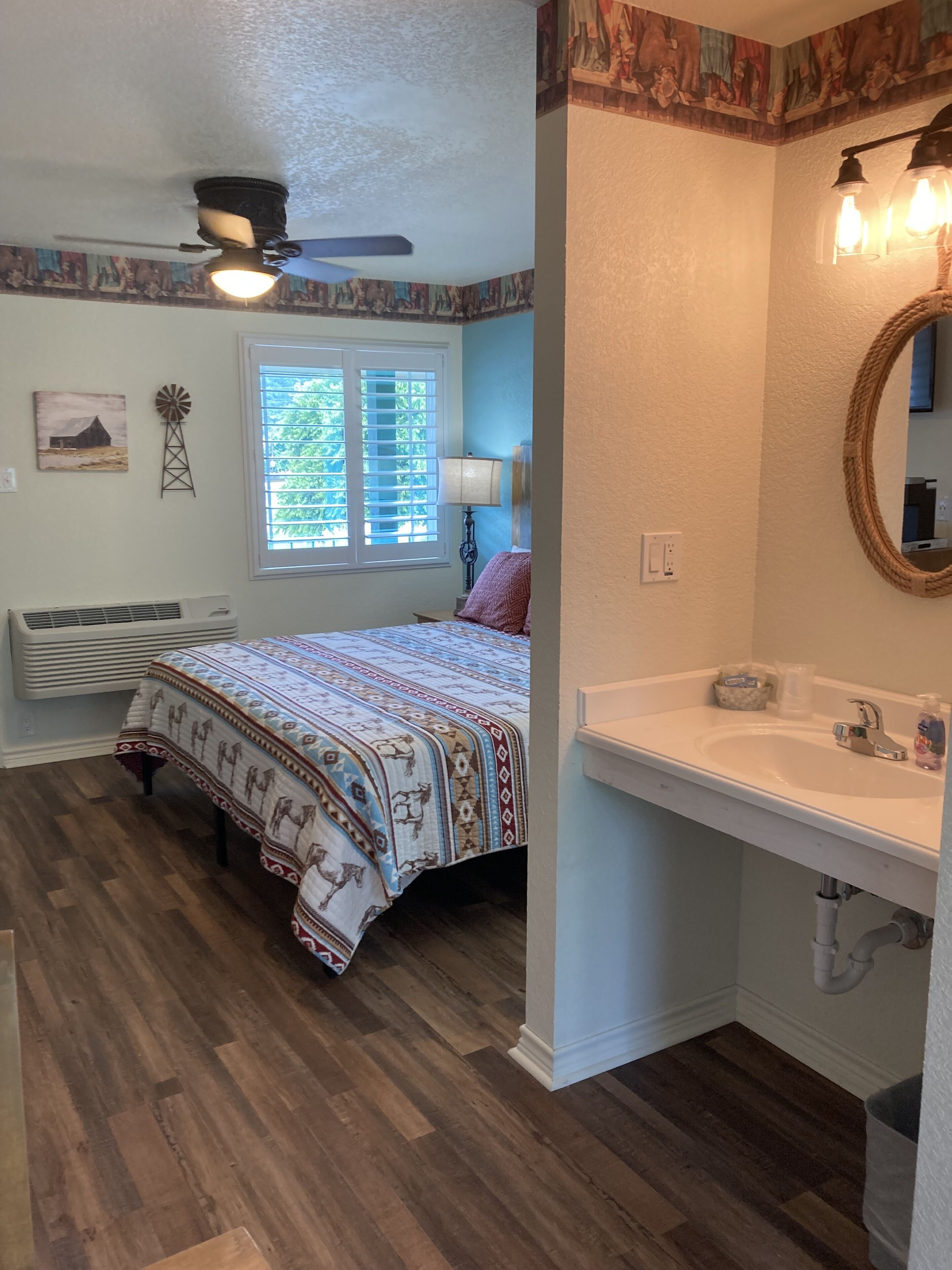 A bedroom with a bed, a ceiling fan, a window with blinds, a wall-mounted air conditioner, and a bathroom with a sink and mirror.