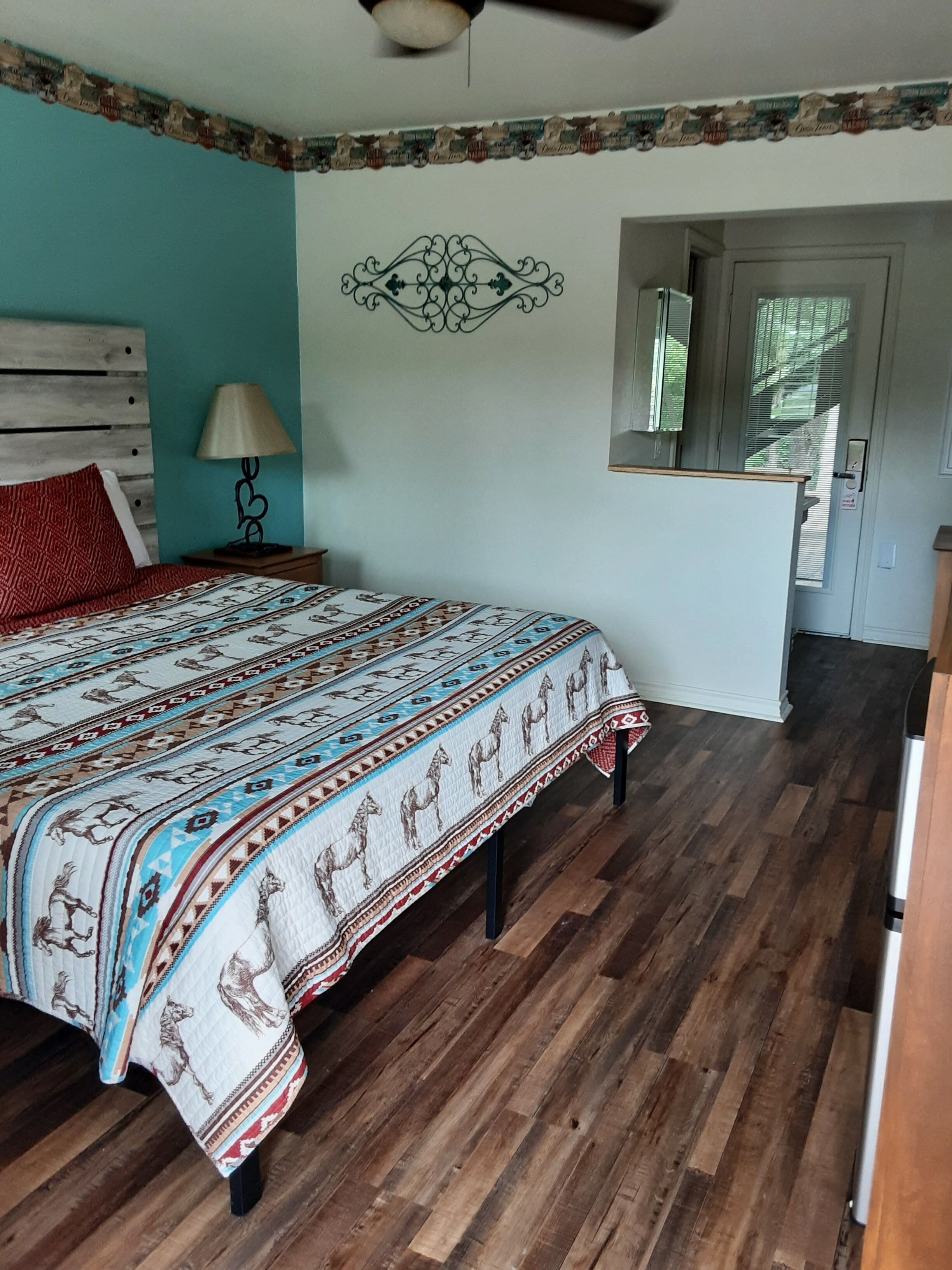 Une chambre à coucher avec un lit en bois, une couvre-lit à motifs, une table de chevet avec une lampe, un élément décoratif mural et une porte dotée d’un miroir.