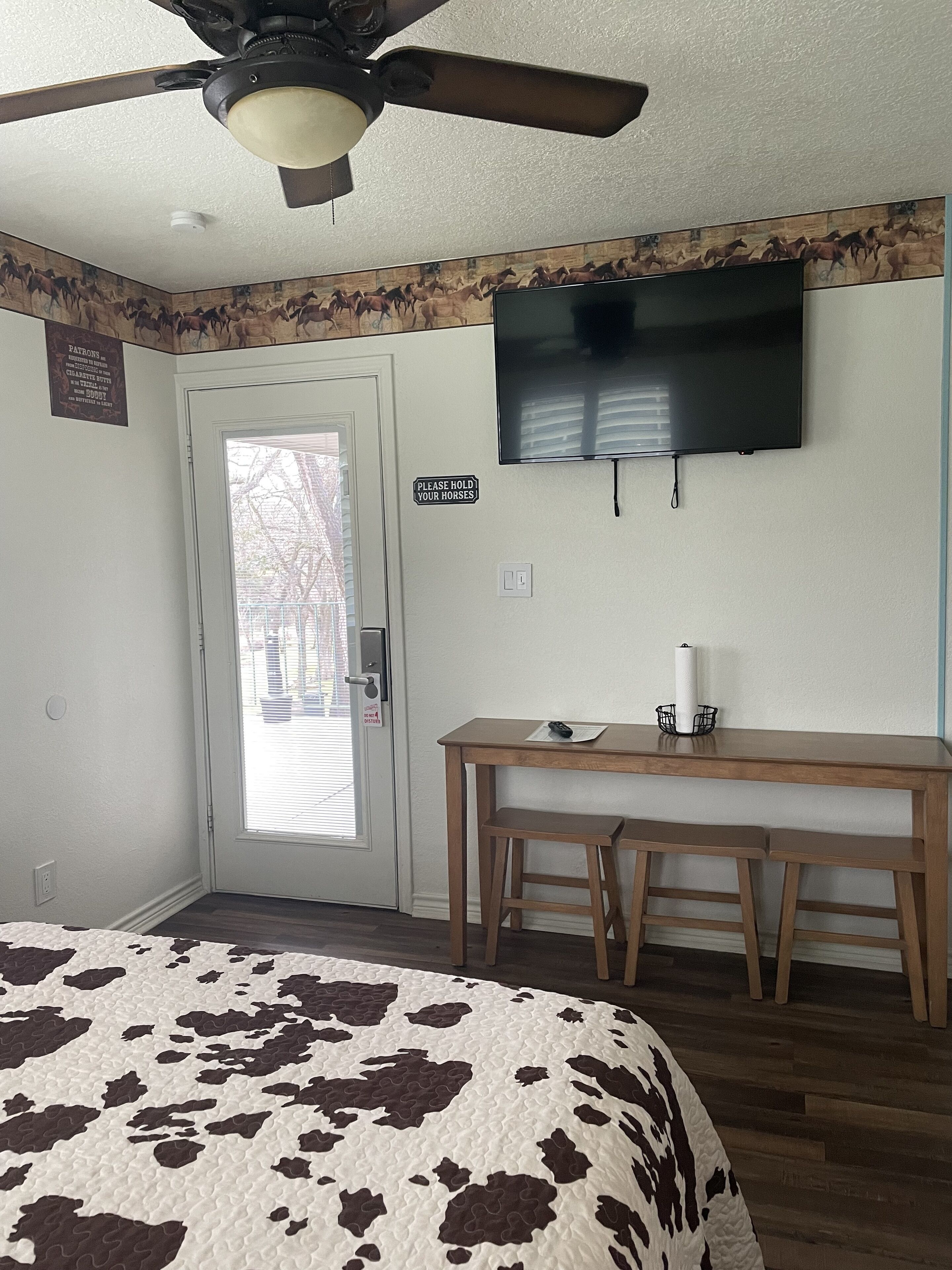 A bedroom with a bed, a desk, a chair, a television, a ceiling fan, and a door with a sign.