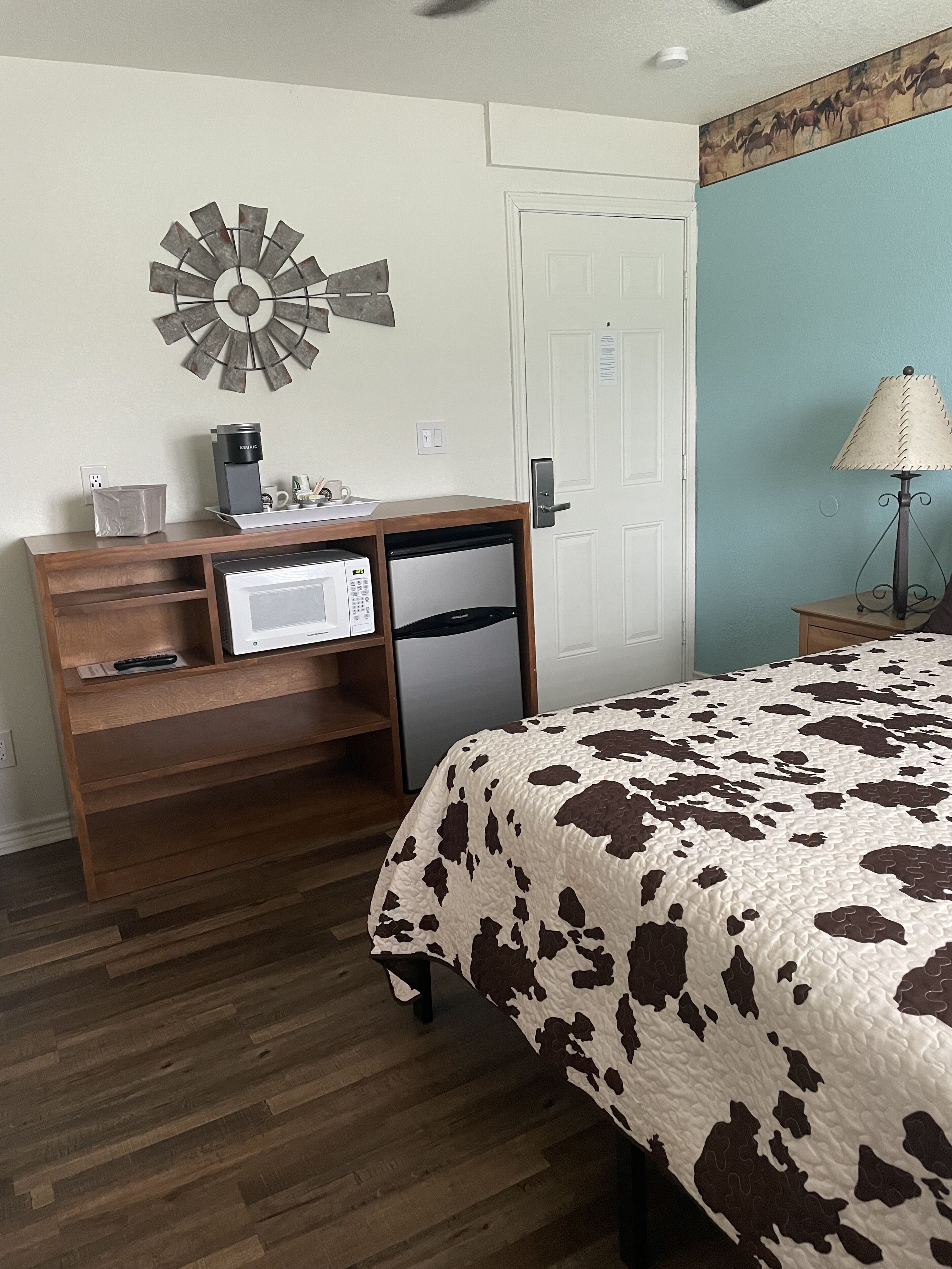 A bedroom with a bed, a wooden desk with a microwave, a small refrigerator, and a wall clock.