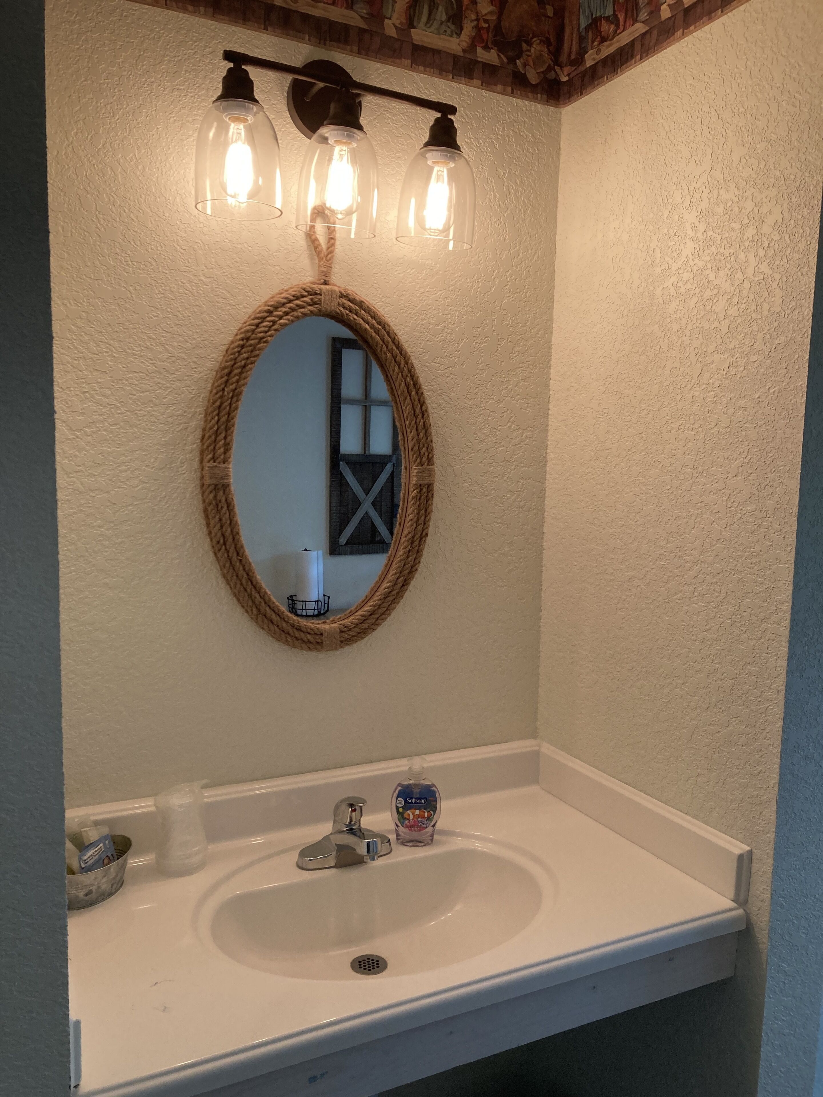 Une salle de bain avec un lavabo blanc, un miroir encadré d’une corde et trois appliques murales.