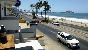 A street view with a white SUV driving on a road lined with palm trees and a beach in the background. - Condo Rusti (Mazatlán)