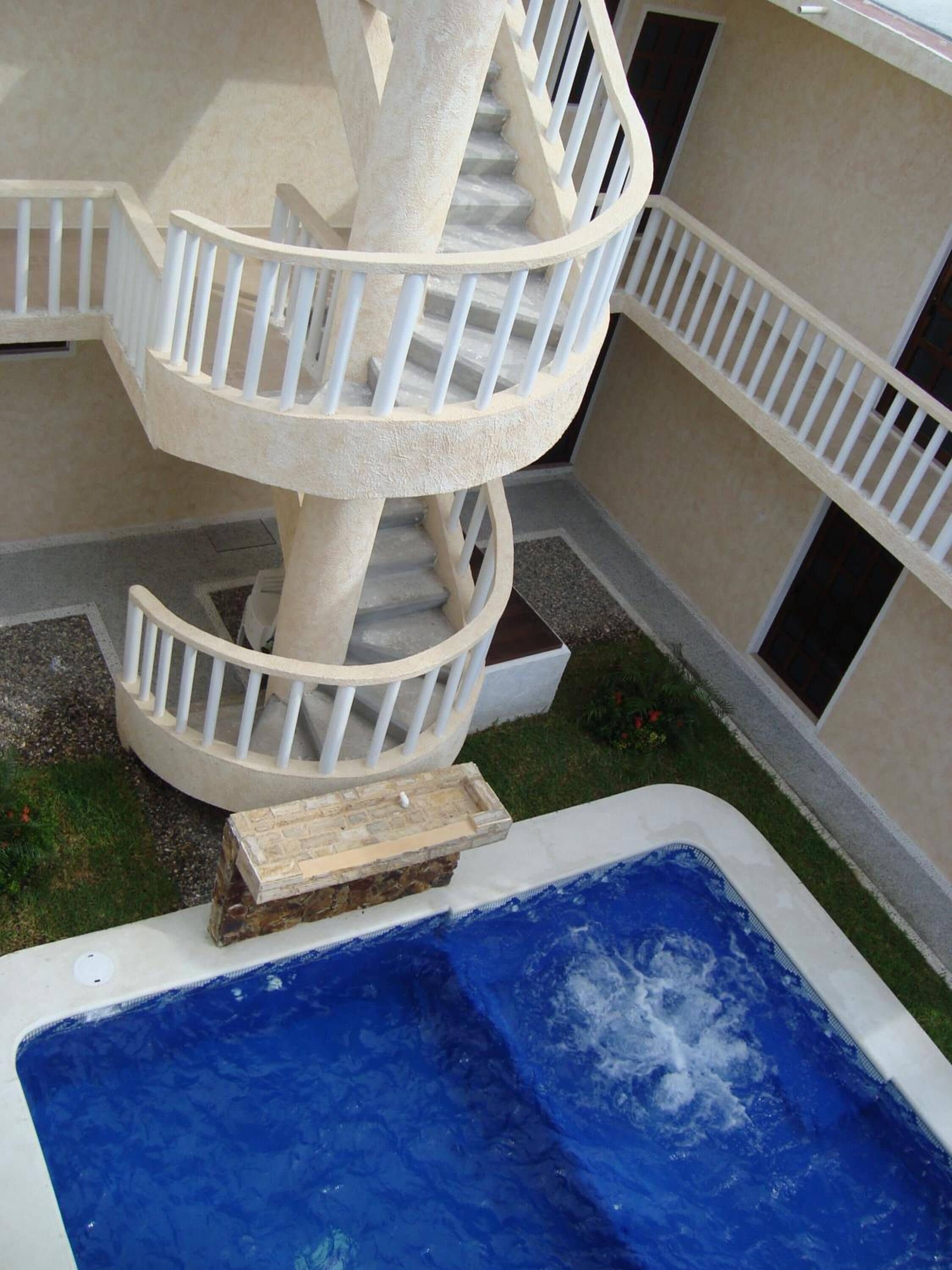 Eine Wendeltreppe mit weißen Geländern führt auf eine höhere Ebene, ein kleiner Pool mit einer Holzterrasse und ein Gebäude mit beigen Wänden und Fenstern.