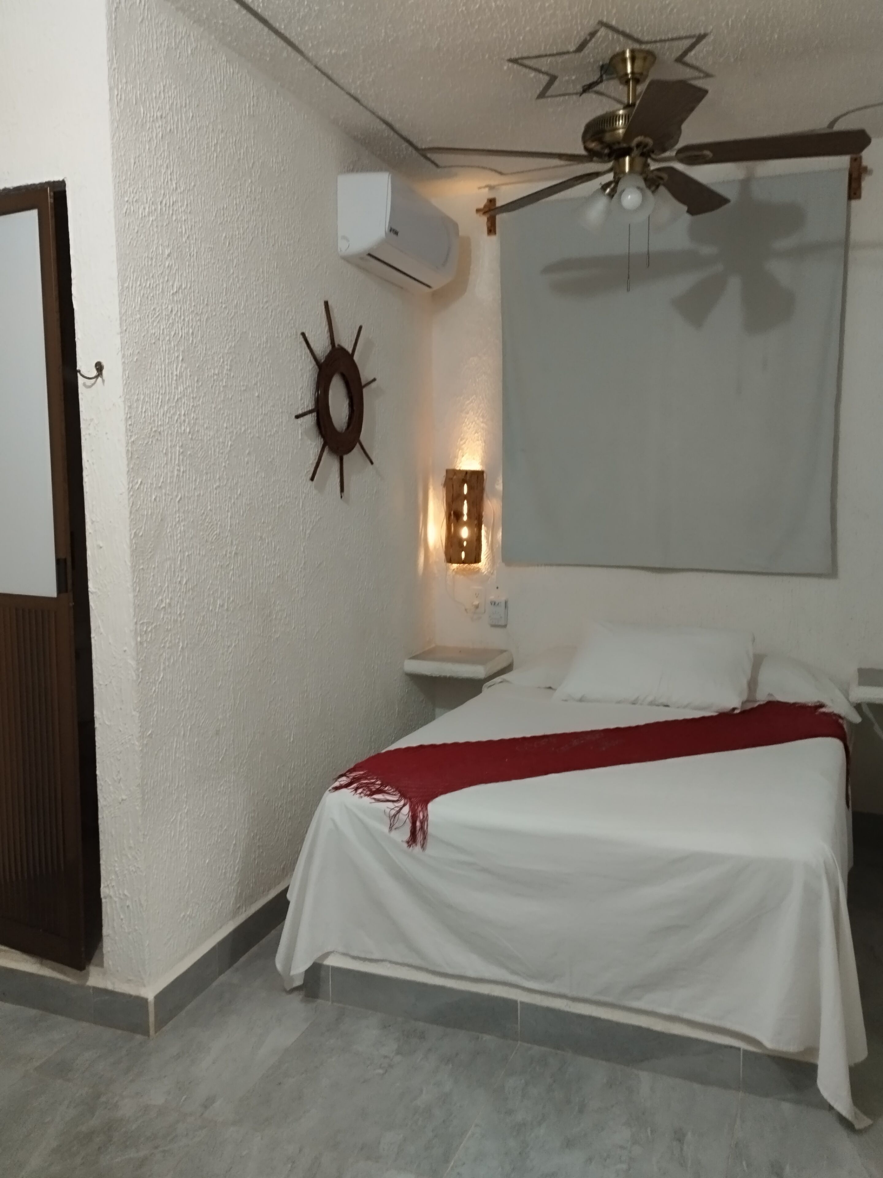 A bedroom with a bed, a ceiling fan, an air conditioning unit, a wall-mounted light, and a decorative sun-shaped object.
