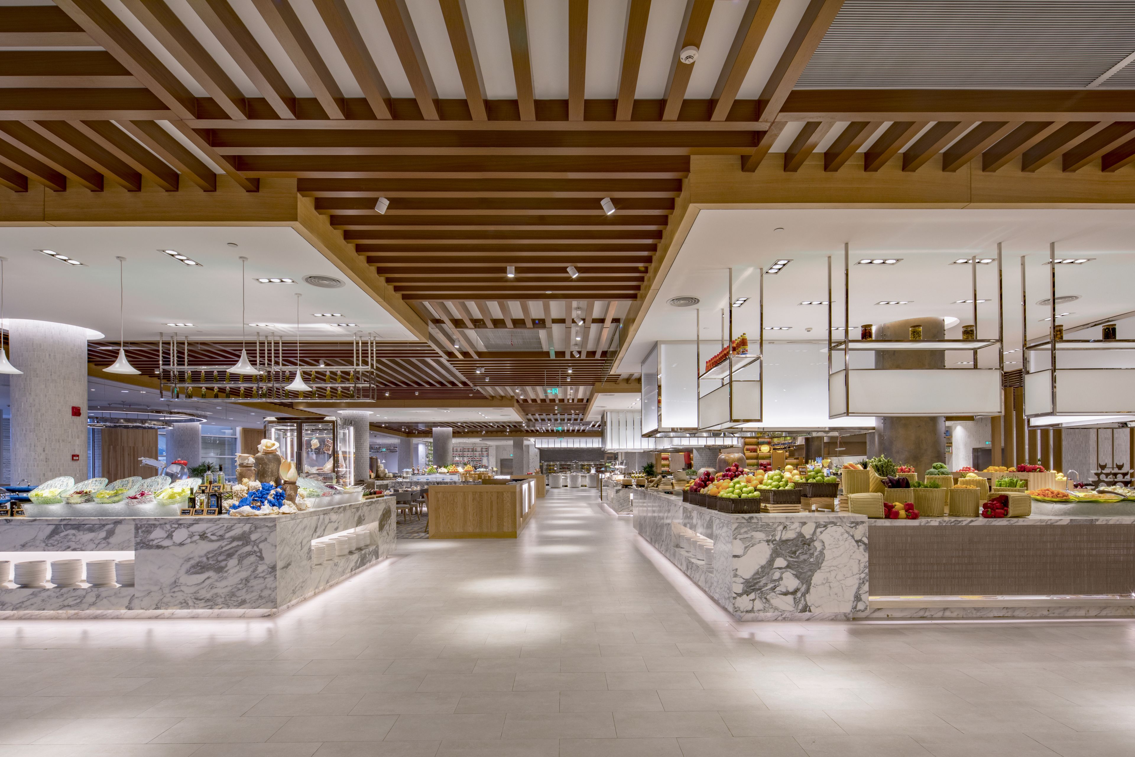 A spacious, modern hotel lobby with marble counters, wooden ceiling beams, and a central island with food displays.