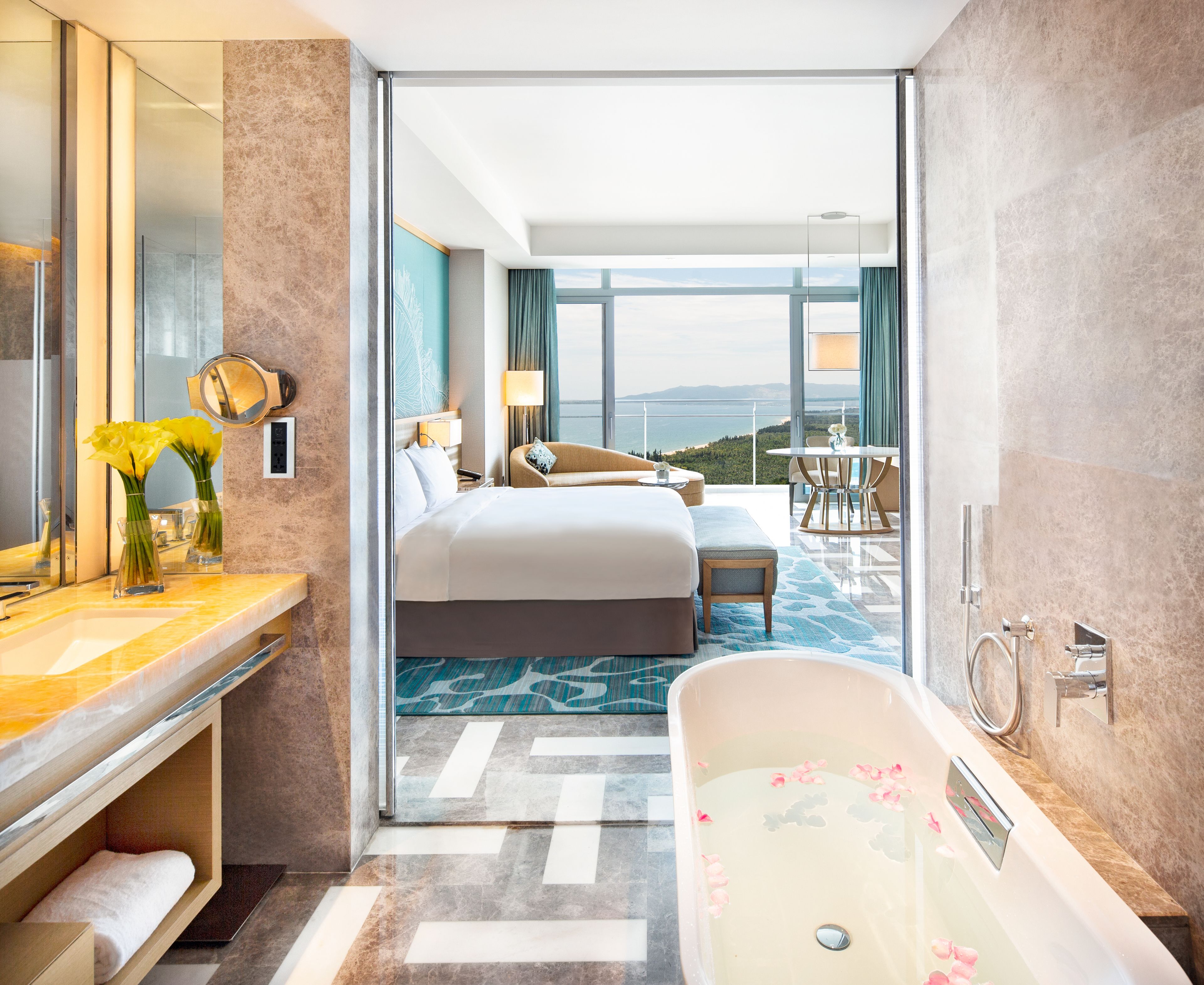 A modern hotel room with a large bed, a sitting area with a view, a bathroom with a bathtub, and a balcony with a view.