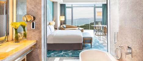 A modern hotel room with a large bed, a sitting area with a view, a bathroom with a bathtub, and a balcony with a view.