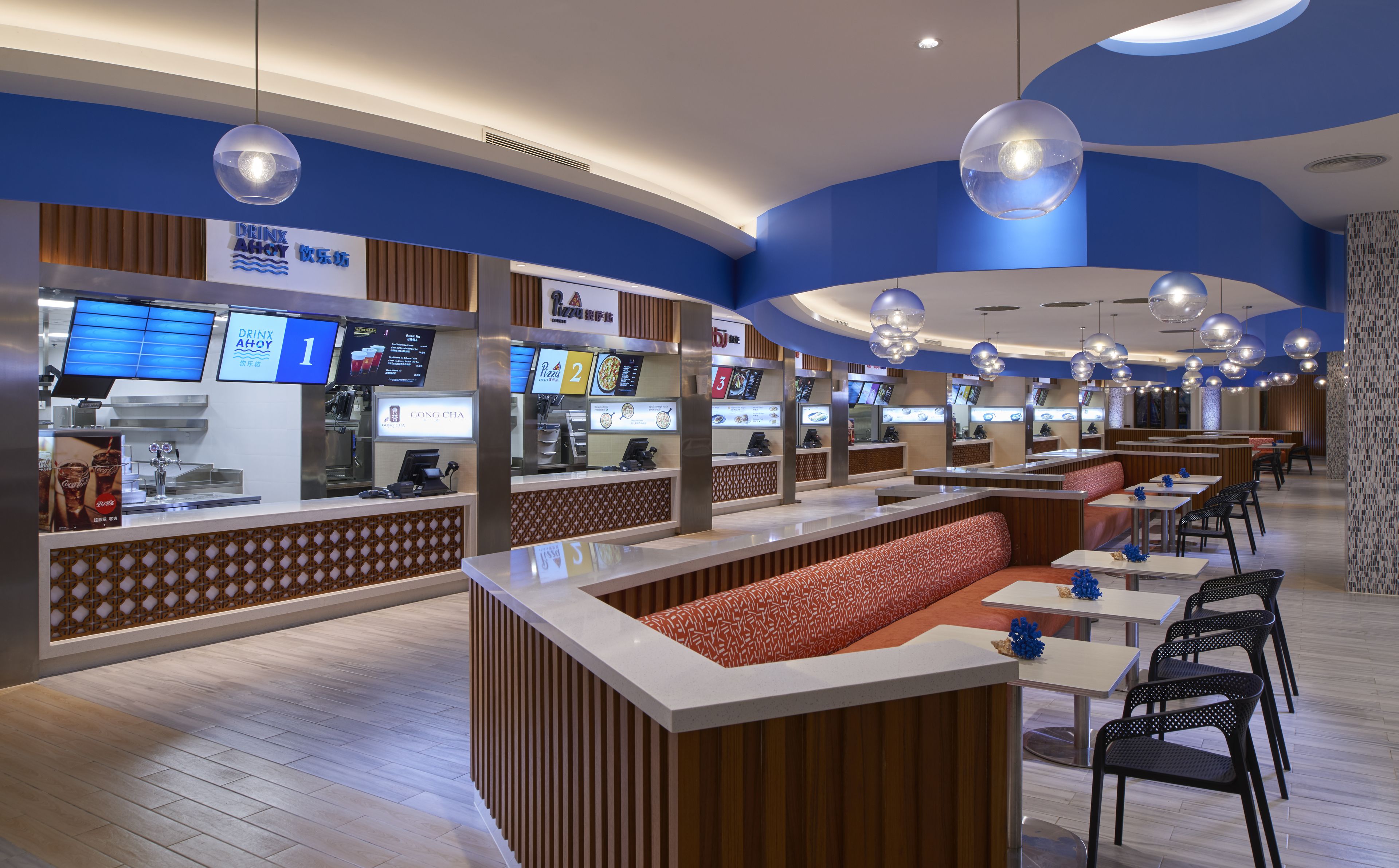 Un restaurante moderno con una decoración azul y blanca, que cuenta con una barra con pantallas digitales del menú, áreas de estar con cojines estampados e iluminación colgante.