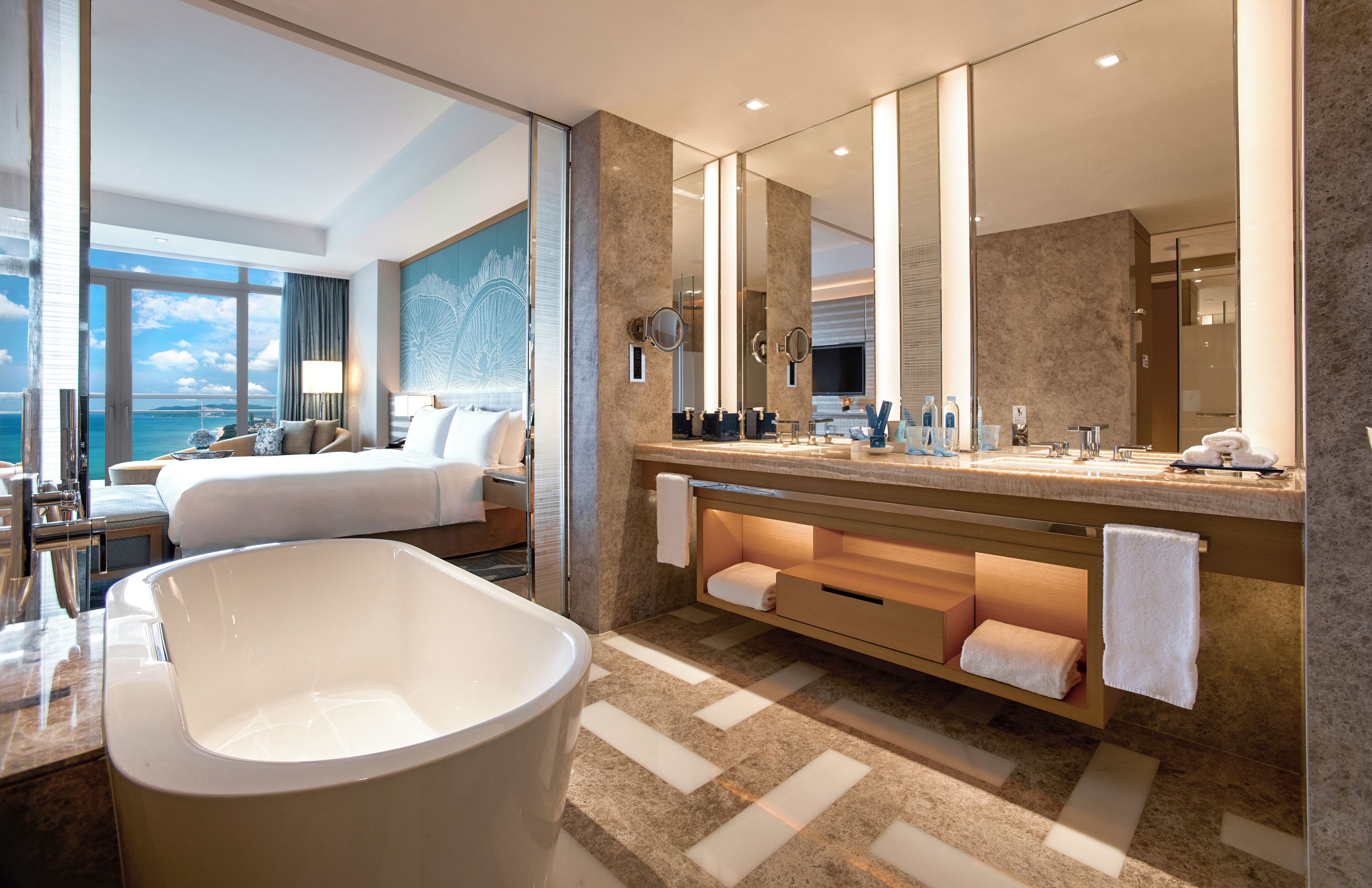 A modern hotel bathroom with a large bathtub, a vanity with a mirror, and a large window offering a view of the ocean.