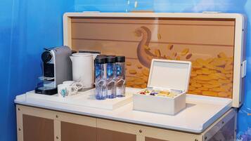 A breakfast setup with a coffee maker, cups, water bottles, and a box of cereal on a counter.