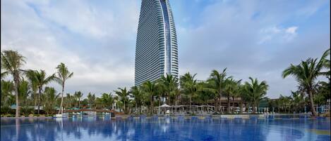 Un hotel moderno con un edificio alto y curvo, una gran piscina con azulejos azules y palmeras alrededor.