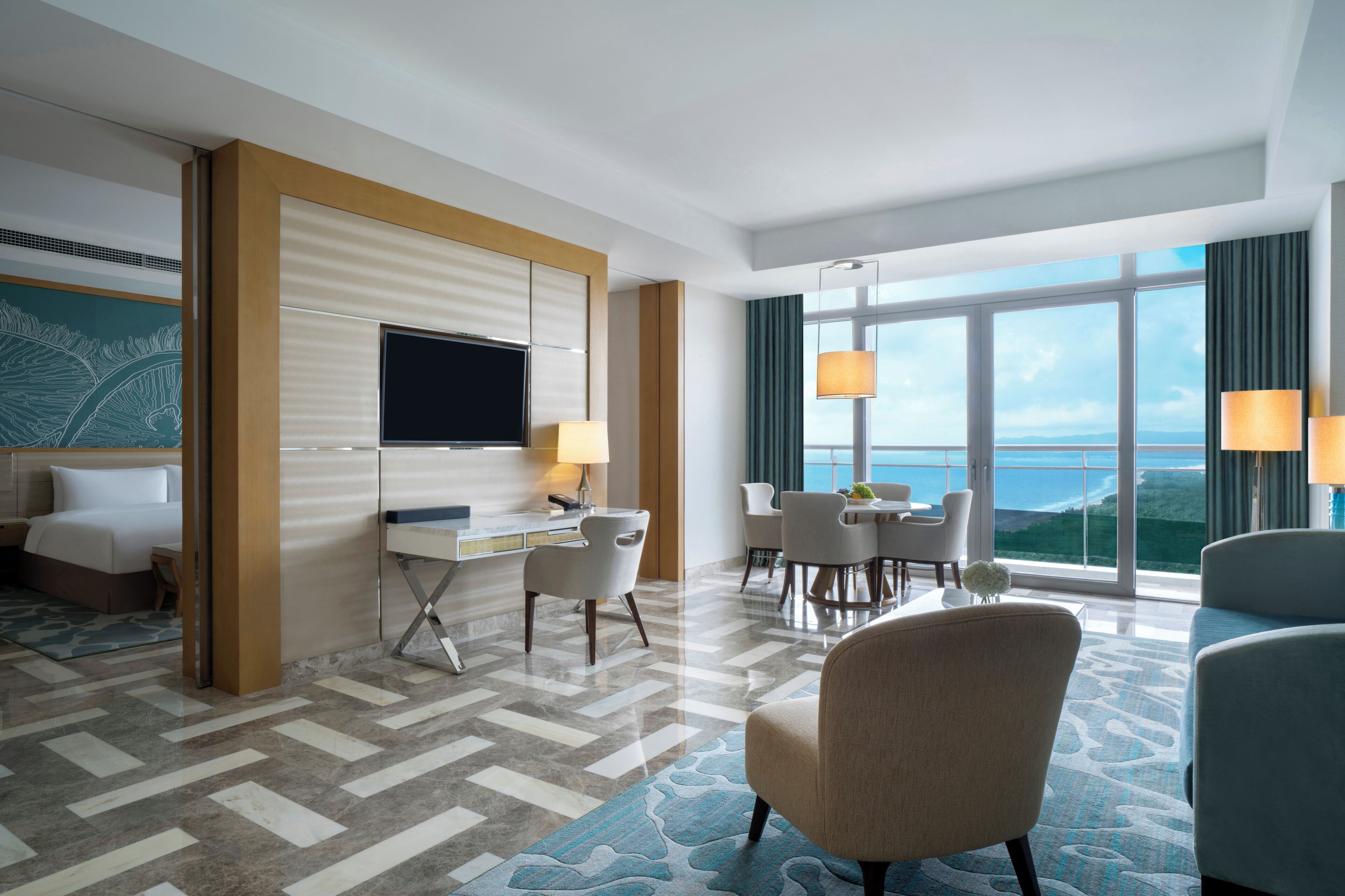 A hotel room with a bed, desk, and chairs. There is a TV on the wall and a large window with a view of the ocean.