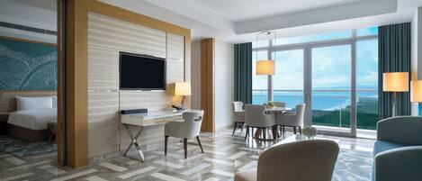 A hotel room with a bed, desk, and chairs. There is a TV on the wall and a large window with a view of the ocean.