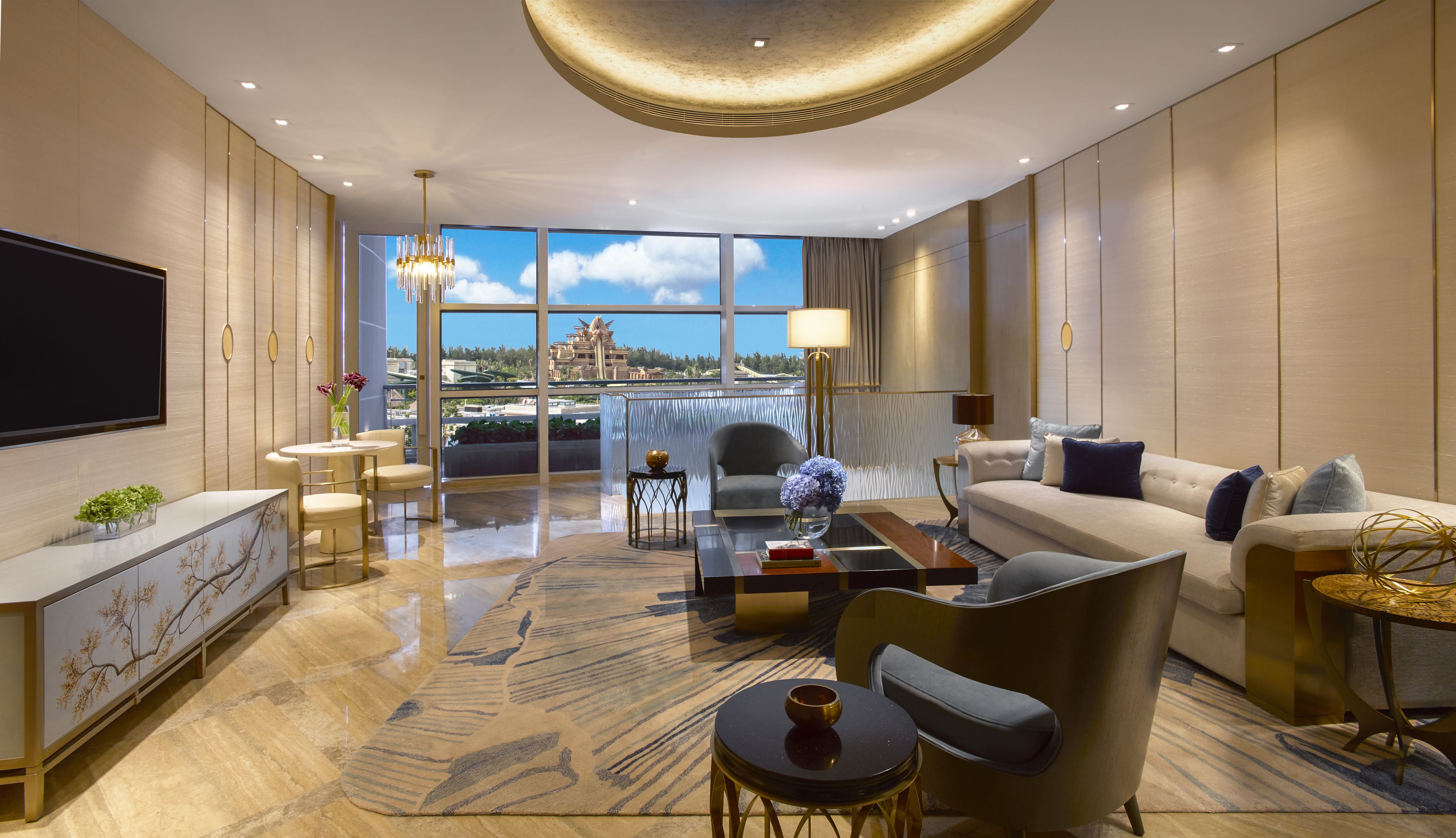 A modern living room with a large TV, comfortable seating, a view of the city, and a balcony with a scenic view.