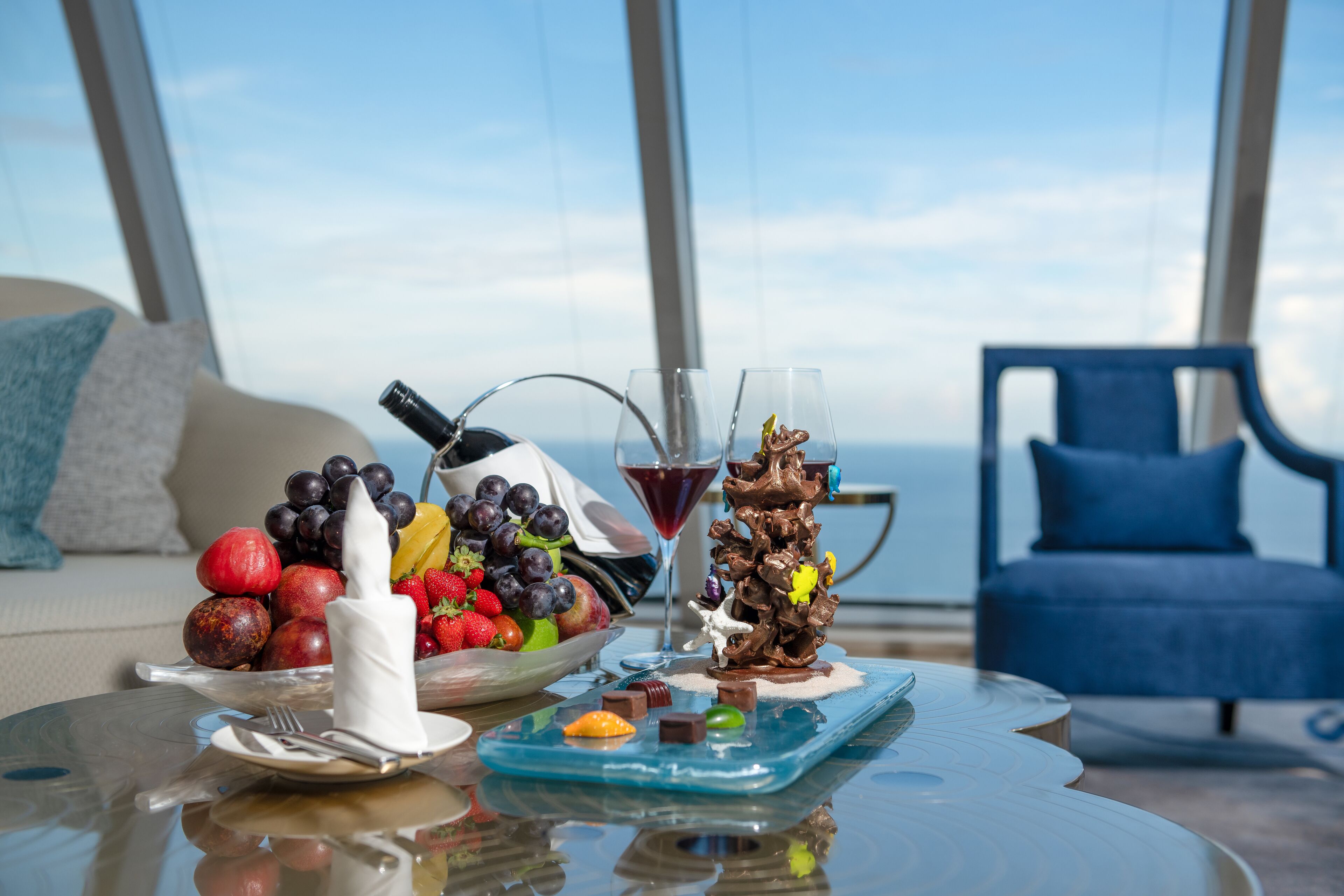 A dining table with a fruit bowl, a chocolate sculpture, and a wine glass on a balcony with a view of the ocean.