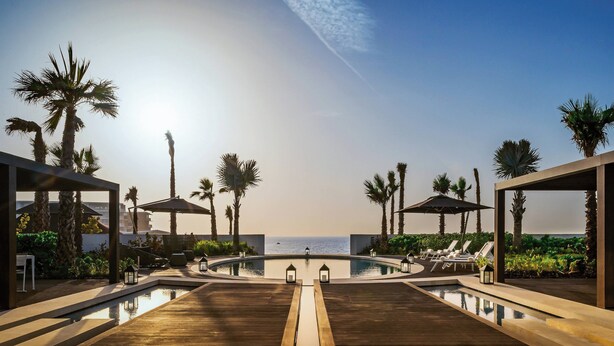 Ein Poolbereich mit Liegestühlen und Sonnenschirmen, Palmen und Blick aufs Meer.
