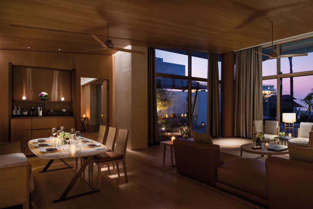 A modern hotel room with a dining area, a sofa, and a large window with a view of the evening sky and palm trees.