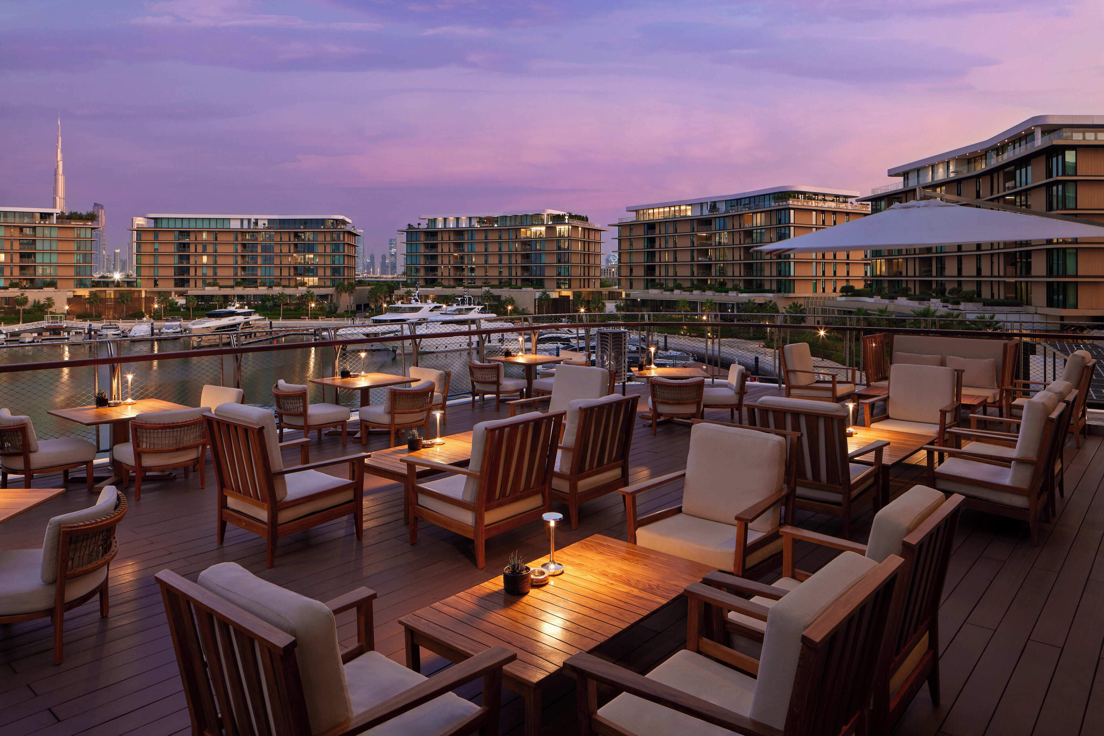 A rooftop bar with outdoor seating, overlooking a marina and modern buildings.