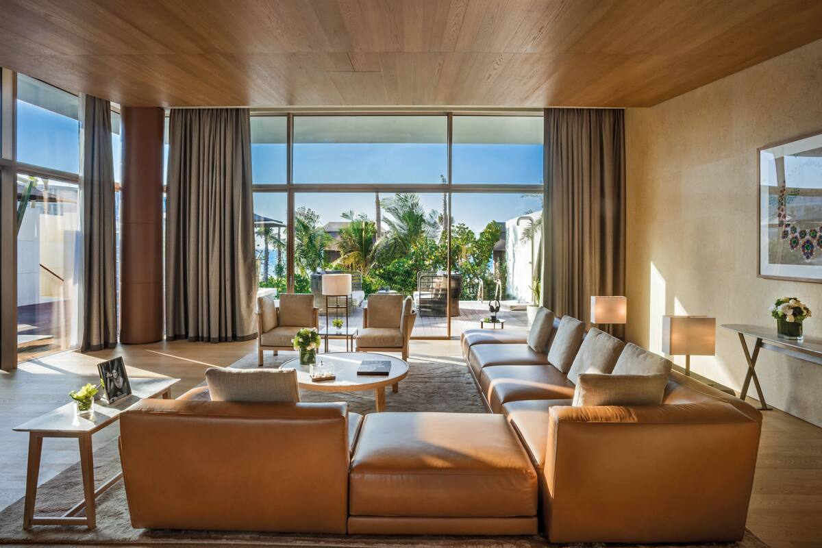 A spacious living room with a large sofa set, a coffee table, and a view of a tropical outdoor area through large windows.