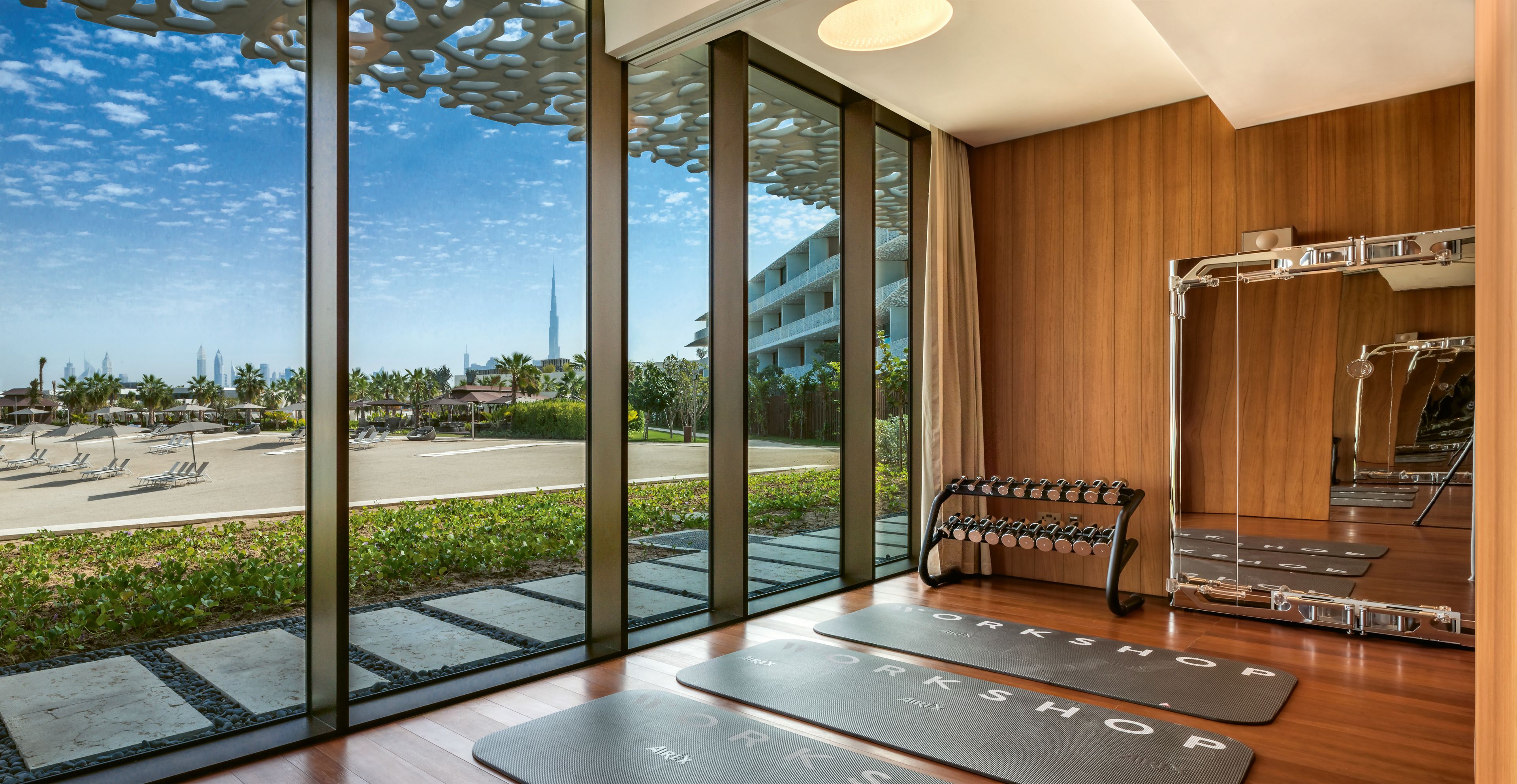 A modern hotel room with large windows offering a view of a cityscape, a wooden floor, and a yoga mat area.