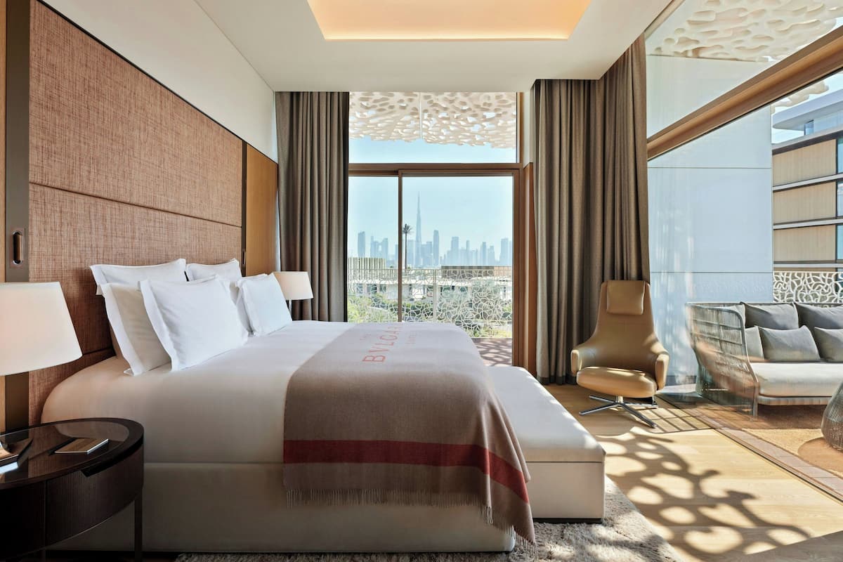 A hotel room with a large bed, a sitting area with a sofa and armchairs, a view of a city skyline through large windows, and a decorative ceiling panel.
